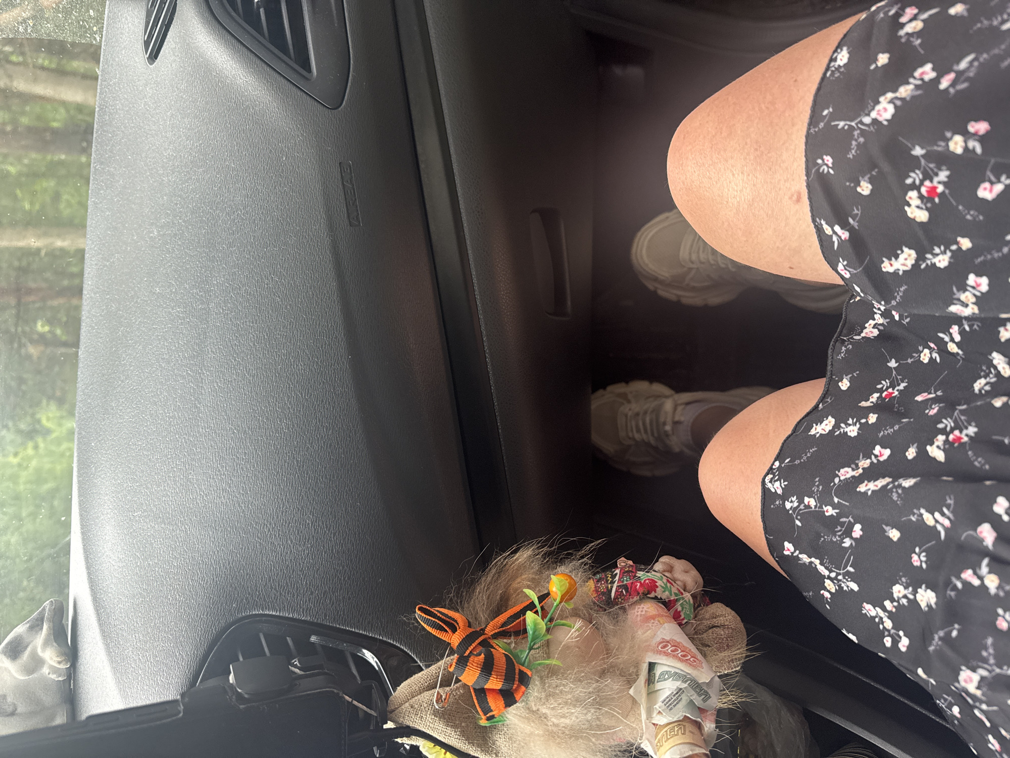 The image shows the interior of a car, looking forward from the passenger seat. The dashboard is visible, as is the leg of the person taking the photo, wearing a black floral dress and light-colored sneakers. To the left of the frame, a stuffed gnome-like figure with a St. George's ribbon and a small orange decoration is hanging from the dashboard, with rolled-up banknotes tucked into its fabric. The view through the windshield shows trees and greenery, suggesting an outdoor or roadside location. The specific location in Stepanovo, Russia is not discernible from the image itself.