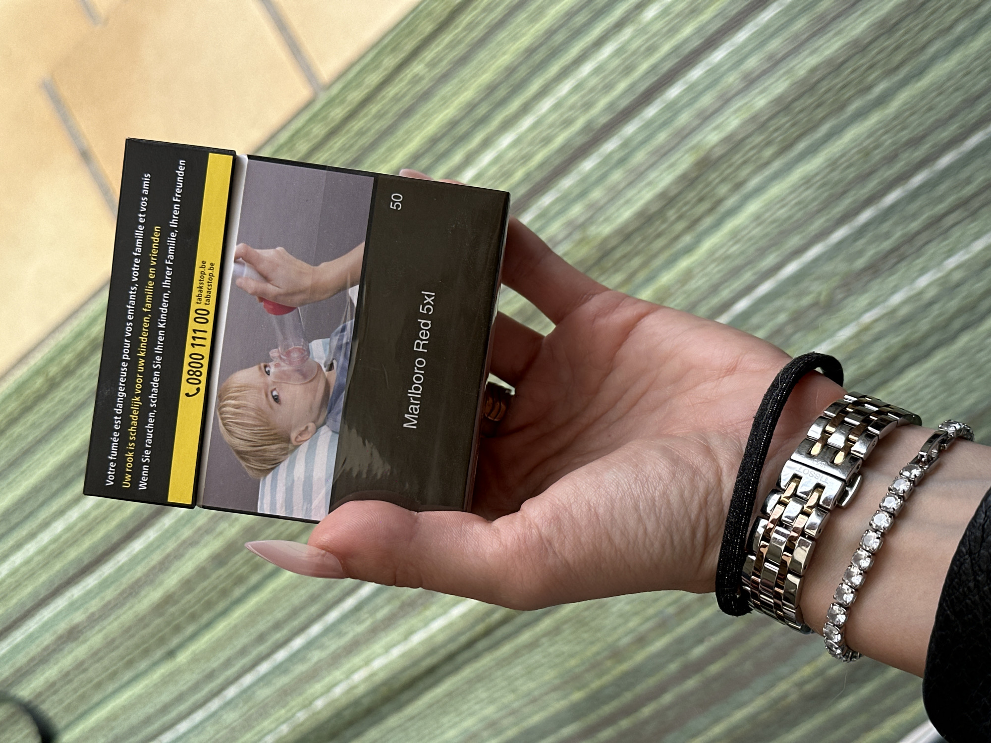 The image displays a person's hand holding a pack of Marlboro Red 5xl cigarettes. The pack has warning text in multiple languages, including French, Dutch, and German, and a graphic image of a child with a pacifier. The background is a green, striped surface. A silver and gold watch and a diamond bracelet are visible on the person's wrist. There is no discernible location in the city of Bruges, Belgium.