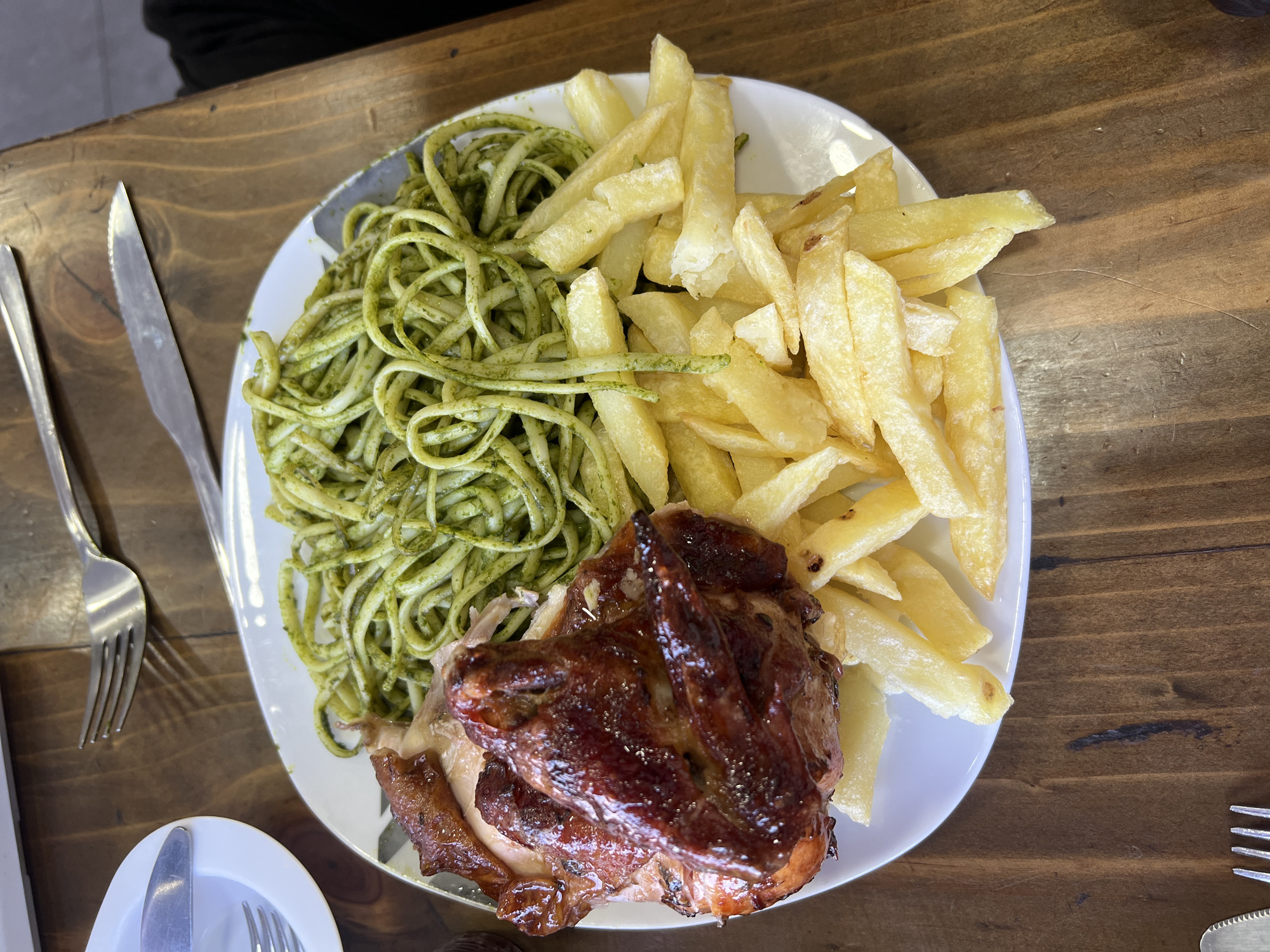 On a wooden table, a white plate holds a meal consisting of green spaghetti, French fries, and roasted chicken. A knife and fork are positioned to the left of the plate. The scene takes place in a restaurant in Arica, Chile, as this meal is typical of the region. The focus is on the plated food, detailing its composition and arrangement. No people are visibly interacting with the food.
