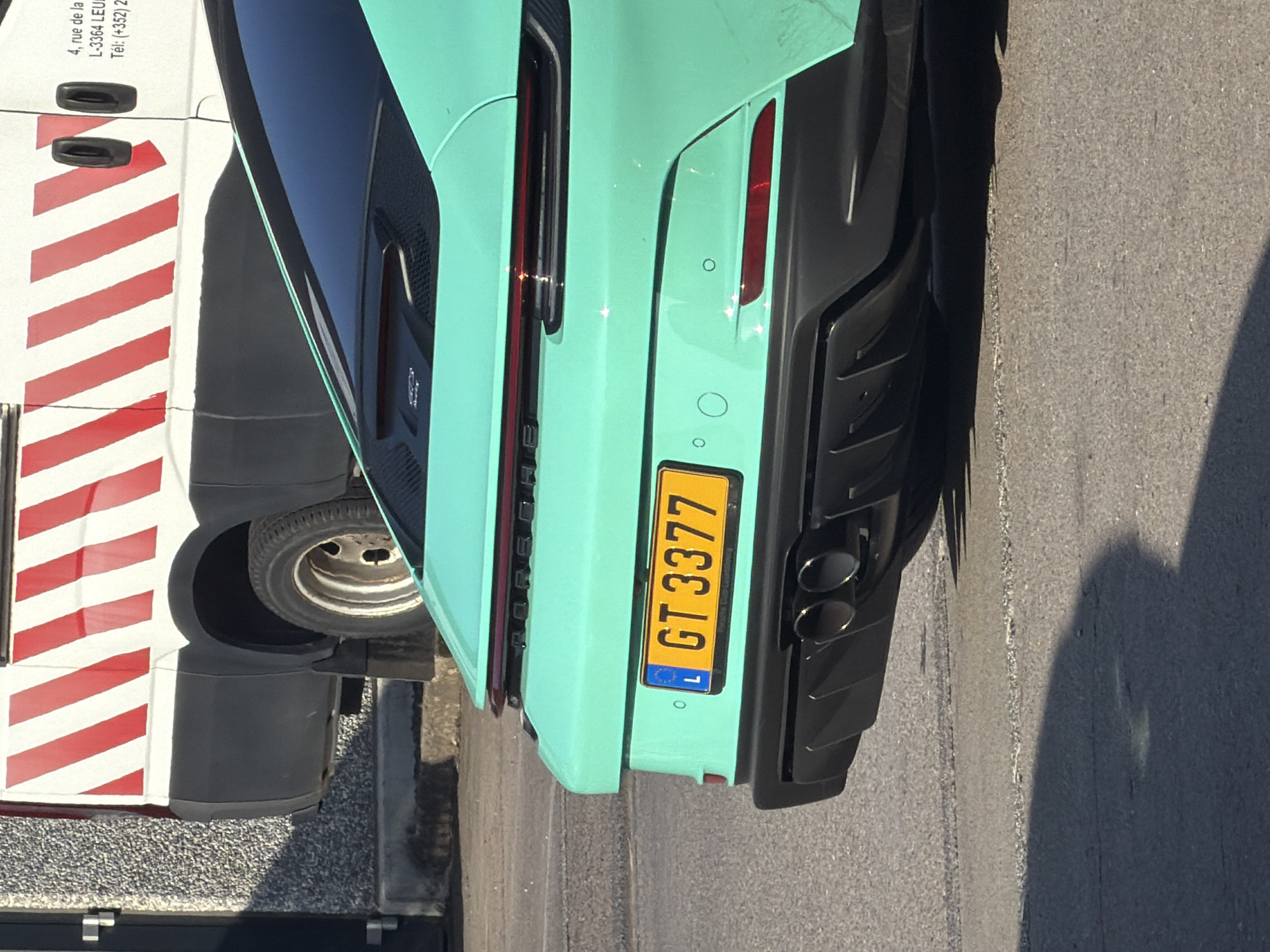 A close-up rear view of a turquoise Porsche 911 is parked on an asphalt road in Leudelange, Luxembourg. The car's license plate reads "GT 3377" and is yellow with a blue European Union symbol on the left. To the left of the Porsche, a white van with red diagonal stripes is partially visible. The van has "4, rue de la L-3364 LEUDELANGE" and "Tél: +(352) 2" printed on its side. The background consists of a grey textured wall and an asphalt ground, casting a shadow of the car. The lighting suggests it is daytime, with the sun casting a long shadow to the right of the vehicle.