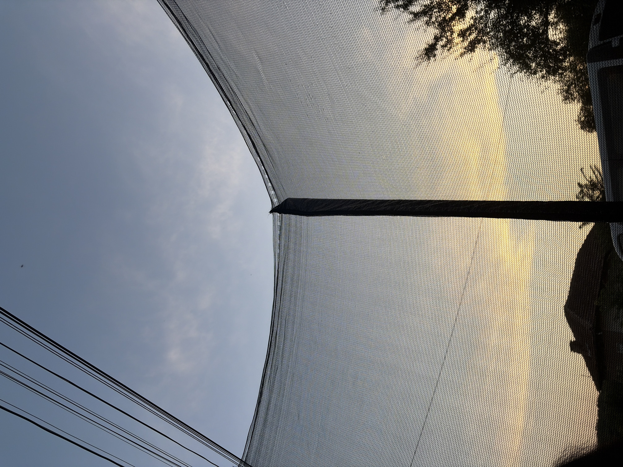The image shows a view of the sky through a trampoline net at sunset. Several power lines are visible in the upper left corner against a pale blue sky. The setting sun casts a warm yellow and orange glow across the horizon, partially obscured by the net. In the lower portion of the image, the silhouette of a roof of a building and trees are visible. The image appears to have been taken from within a residential area in Karamanovo, Bulgaria, as suggested by the presence of the house and trees.