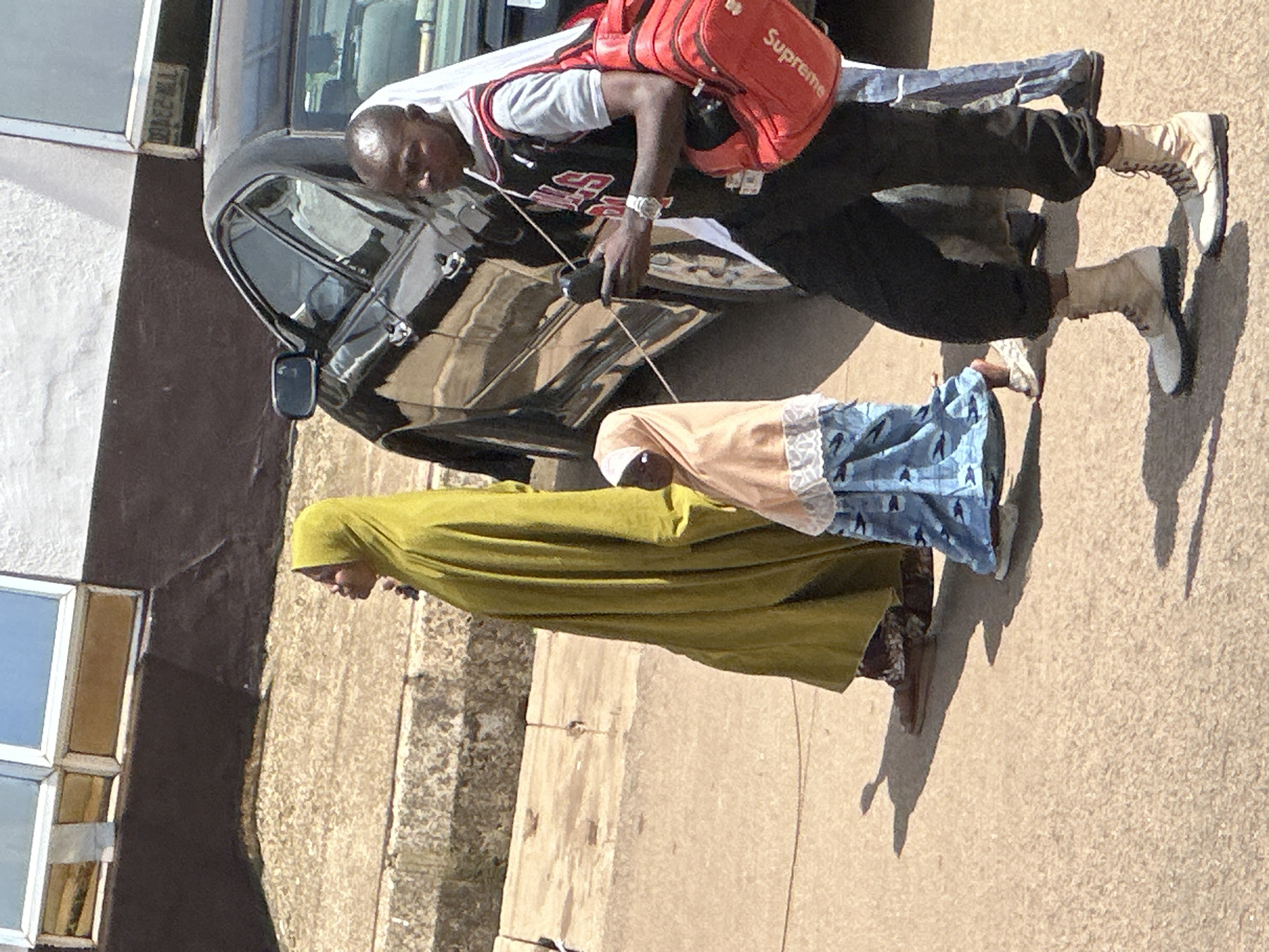 In Bukuru, Nigeria, three individuals walk on a paved surface beside a black car. The foremost person, a male, wears a grey t-shirt, a black and red basketball-style jersey, black trousers, and light-colored combat boots. He carries a red duffel bag with "Supreme" written on it over his right shoulder and holds a black object in his right hand. To his left, a young child, dressed in a light brown hijab and a blue patterned skirt, walks. Behind the child and slightly to the left, an adult, dressed in a flowing olive-green garment and a headscarf, walks with their head turned to the left. The background features a concrete wall with windows and a section of the black car, indicating an urban setting.