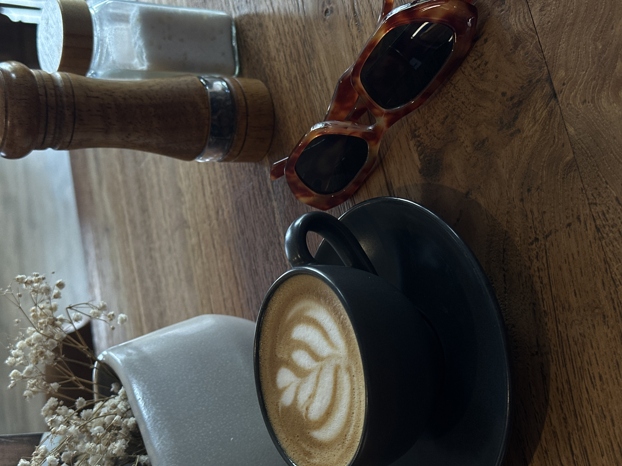 This media file captures an overhead view of various items arranged on a wooden table, likely in a cafe or home setting in North Kuta, Indonesia. The dominant subject is a black ceramic coffee cup with a saucer, containing a latte beautifully adorned with white foamy latte art in a leaf pattern.
Adjacent to the cup, a pair of stylish tortoise-shell sunglasses with dark lenses rests on the table, suggesting a pause or break. Further up and to the left, a wooden pepper grinder and a glass salt shaker with a wooden lid are visible, indicating a dining context. In the bottom-left corner, a light grey or off-white vase holds a small bunch of delicate, dried white flowers (likely baby's breath), adding a touch of natural elegance to the scene.
The wooden table surface features a distinct grain, varying in shade from medium to dark brown, and appears well-worn in areas, contributing to a cozy and rustic aesthetic. Natural light illuminates the scene, suggesting it is daytime, possibly morning or afternoon. The overall impression is one of relaxation and calm, characteristic of enjoying a quiet moment with a beverage. No people are visible, and there is no discernible text on any of the objects.
