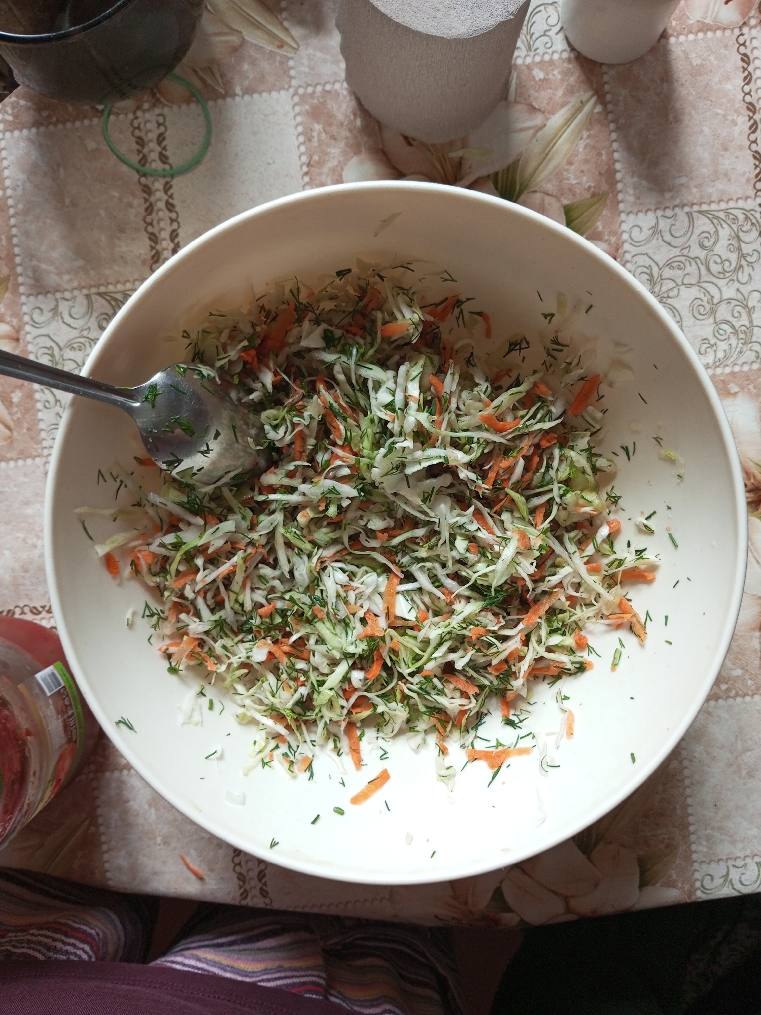 The image shows a close-up, high-angle shot of a white bowl filled with a shredded cabbage and carrot salad, mixed with dill. A metal spoon rests in the salad. The bowl is positioned on a table covered with a patterned tablecloth featuring floral motifs and a beige and brown checkered design. In the background, a dark pot or container is partially visible on the left, and a roll of what appears to be paper towel or toilet paper stands on the right. The overall scene is indoors, likely a kitchen or dining area, but no specific location in Ароматновское сельское поселение, Ukraine is discernible. There are no people visible in the image.