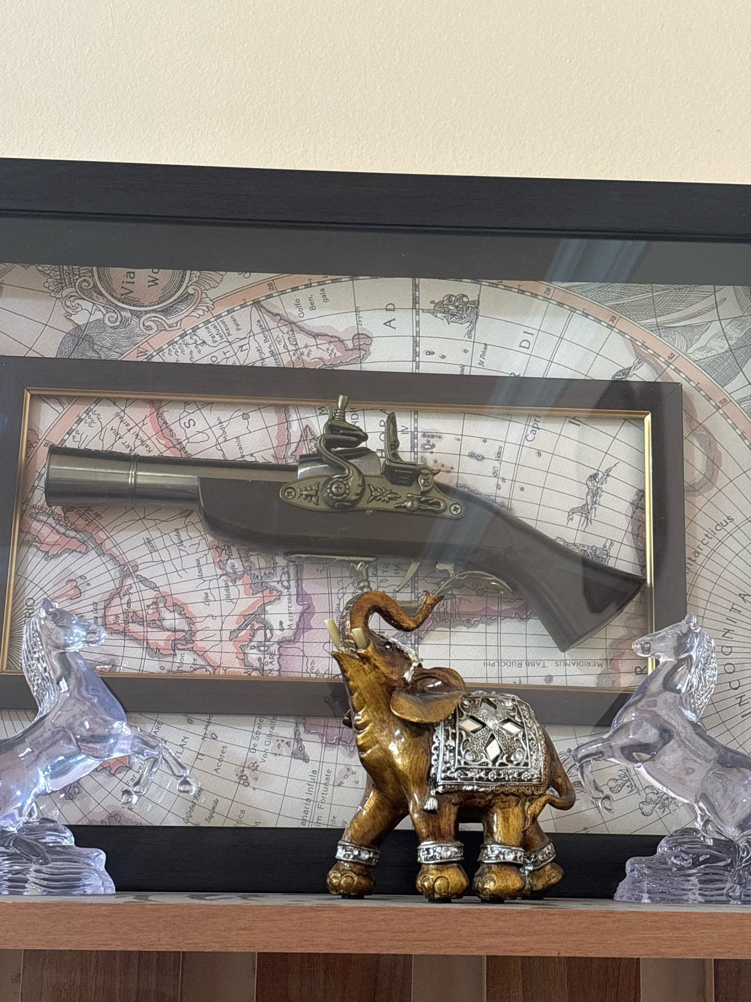 The image shows a framed antique flintlock pistol displayed over a vintage map. The pistol is dark brown and metallic, with intricate detailing on the lock mechanism. The map is sepia-toned with grid lines and place names. In front of the framed display, on a wooden shelf, are three decorative items: a golden elephant figurine adorned with silver and rhinestone details, and two clear acrylic horses in rearing poses. The elephant is centrally positioned, with a horse on either side. The background is a plain, light-colored wall. The scene is indoors, likely a home or display setting. There is no indication of Ijevan, Armenia in the image.