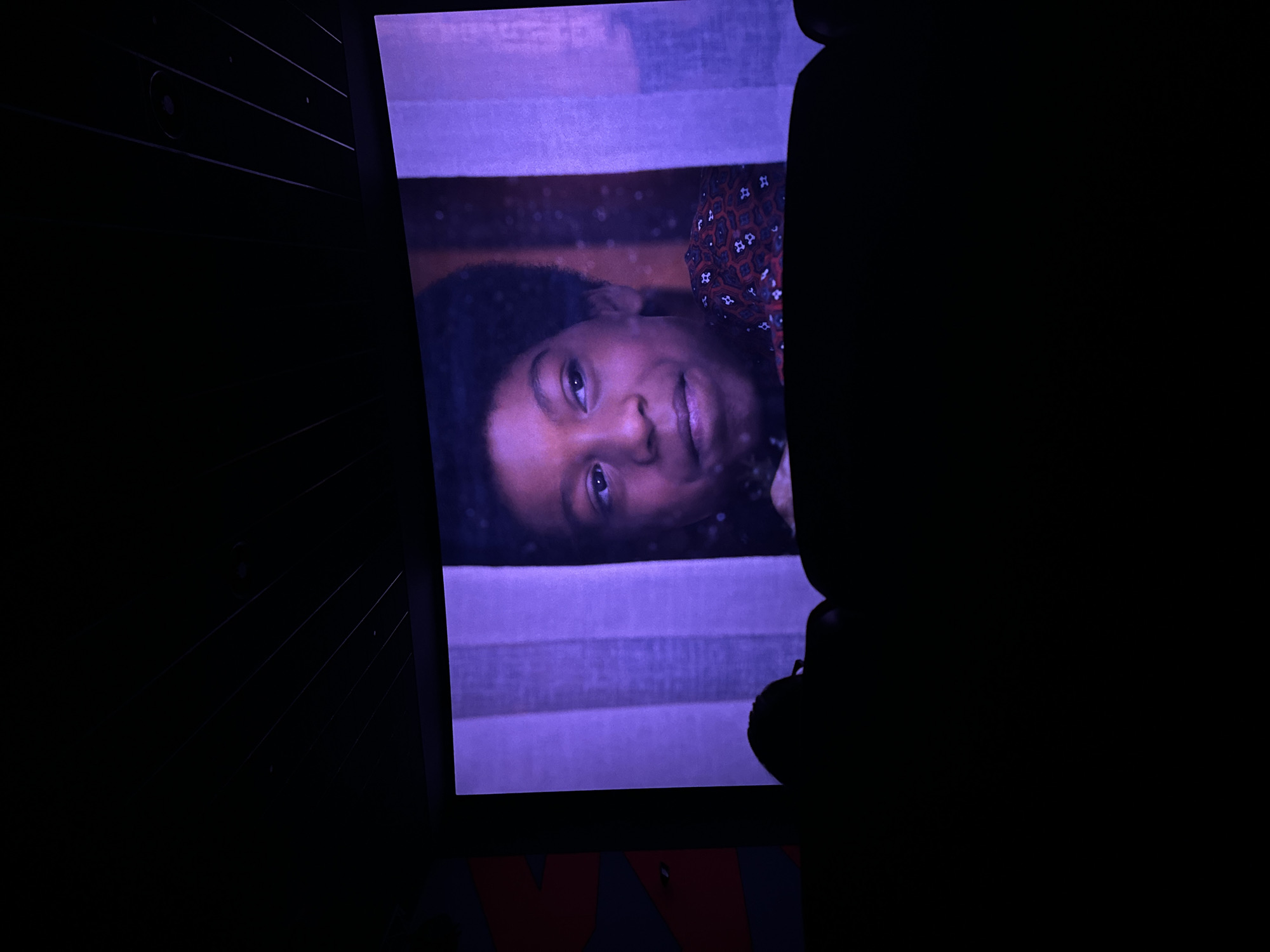 A low-angle, dimly lit shot shows a screen displaying a person with dark hair and eyes, and a patterned shirt. The screen has horizontal lines across it. A dark silhouette of people is partially visible on the right side of the screen. The screen is positioned in front of a dark background with some lines, and the bottom of the screen is above a surface with a red and gray pattern. The location and city are unknown.