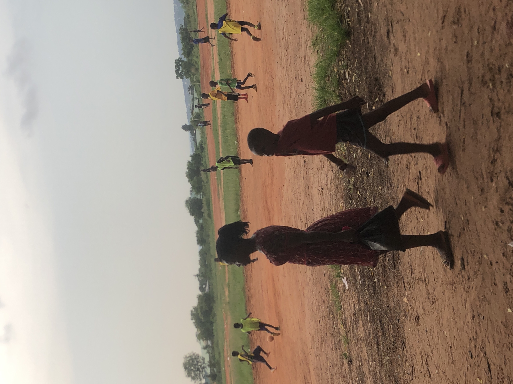 The image depicts an outdoor scene, potentially a field or open space, in Ukwu-Abwa, Nigeria. The ground appears to be a reddish-brown dirt or clay. Several people, possibly children or young adults, are playing soccer in the background. Two other individuals, potentially children, are walking across the foreground. The lighting suggests it might be late afternoon or early morning. The weather is sunny. The sky is a light grey or pale blue. There is some vegetation, including grass and trees, along the edges of the field.