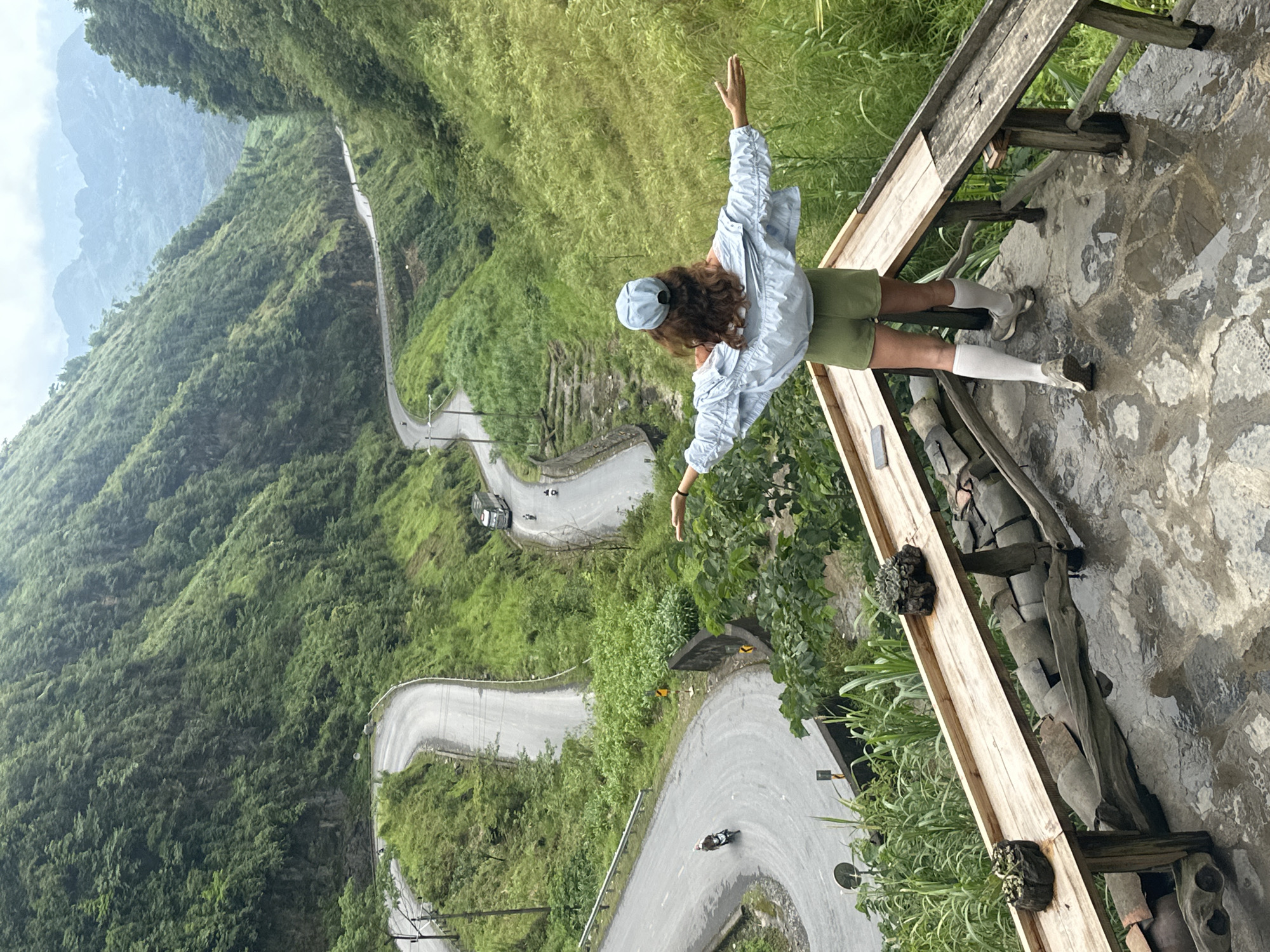 A bird's eye view shows a person standing on a stone patio with a wooden railing in Phố Bảng Commune, Vietnam. The person, presumably a woman with curly brown hair, wears a light blue hat, light blue long-sleeved button-down shirt, green shorts, white socks, and grey sneakers. She is standing with her arms outstretched, facing away from the camera. A long, winding road is visible below, cutting through a lush green landscape. Several vehicles are on the road, including a bus and motorbikes. Mountains are visible in the distance. The patio is paved with stone and has a rustic wooden railing. The lighting suggests it is a bright day, possibly overcast. The scene conveys a sense of freedom and awe in a mountainous landscape.