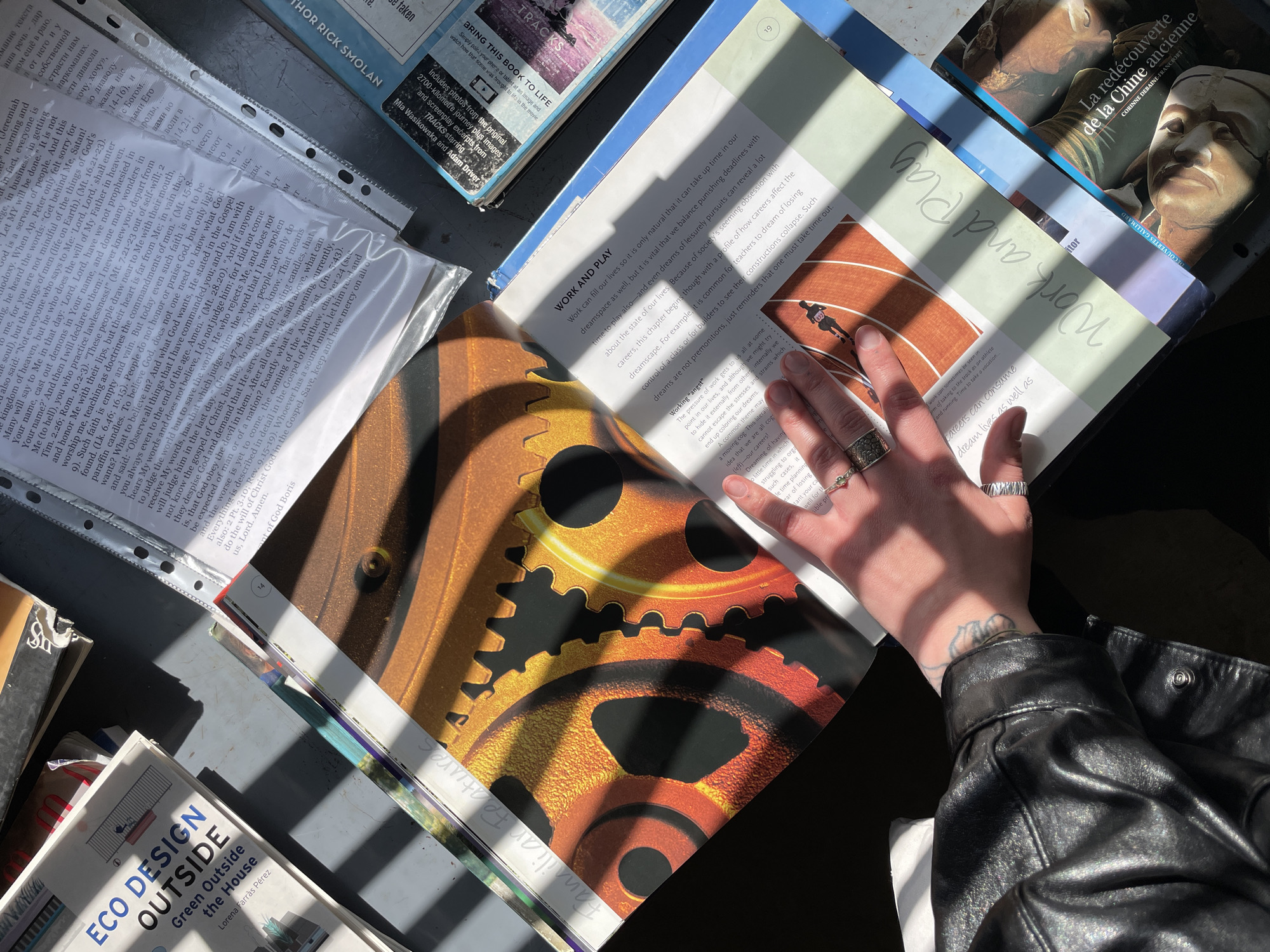 A high-angle shot captures a person's hand resting on an open book amidst a collection of books and papers, bathed in bright sunlight creating distinct shadows. The scene suggests a moment of reading or study, likely indoors in Tbilisi, Georgia, during a sunny day.
The central focus is an open book. The left page features a large, detailed image of interconnected gears in warm orange and gold tones, with "Familiar Features" handwritten near the bottom edge. The right page is titled "WORK AND PLAY" and discusses the balance between work and leisure, with sections like "Working 'angst'" and "Careers can consume dream lives as well as." An image of a running track with a figure is also visible on this page.
Around the open book, several other items are scattered. To the left, papers with extensive text, including religious content with biblical references (e.g., Jeremiah, Matthew, John) and some Cyrillic script, are held in clear plastic sleeves. Below these, a book titled "ECO DESIGN OUTSIDE: Green Outside the House" by Lorena Farrás Pérez is partially visible. To the upper right, two more books are stacked: one with "La redécouverte de la Chine ancienne" by Corinne Debaine-Francfort on its cover, and another blue-spined book mentioning "AUTHOR RICK SMOLAN," "TRACKS," and "Mia Wasikowska and Adam Driver."
The person's left hand, adorned with several rings on the fingers and a small tattoo on the wrist, gently touches the open book. A black leather jacket sleeve is visible in the bottom right corner, suggesting the person is wearing it. The strong sunlight casts striped shadows across the books and papers, highlighting the textures and adding a dynamic quality to the composition.