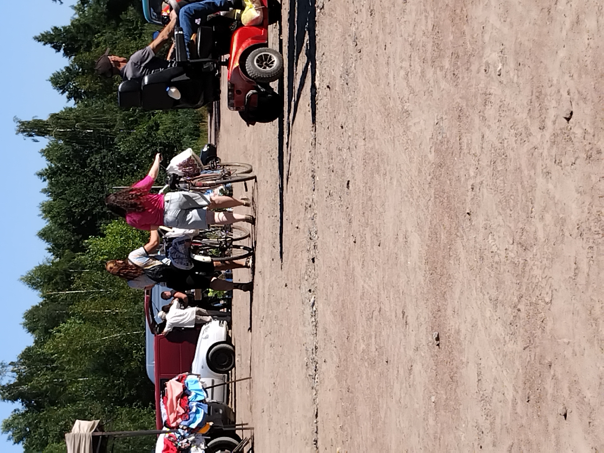In Irshansk, Ukraine, on a sunny day, an outdoor market scene unfolds. The foreground is dominated by a dusty, unpaved ground. In the mid-ground, two women are walking with bicycles. One woman, with long brown hair, wears a plaid shirt and shorts, with a backpack. The other woman, with long dark curly hair, wears a pink shirt and denim shorts. To the right, a man sits in a red mobility scooter, wearing a gray shirt, blue jeans, and a dark baseball cap. He is looking to his right, and a yellow bag is attached to the scooter. Behind the women, a white car is parked next to a red van with its rear doors open. Further back, a tent structure with bundled textiles is visible. The background is filled with a dense line of green trees under a clear blue sky.