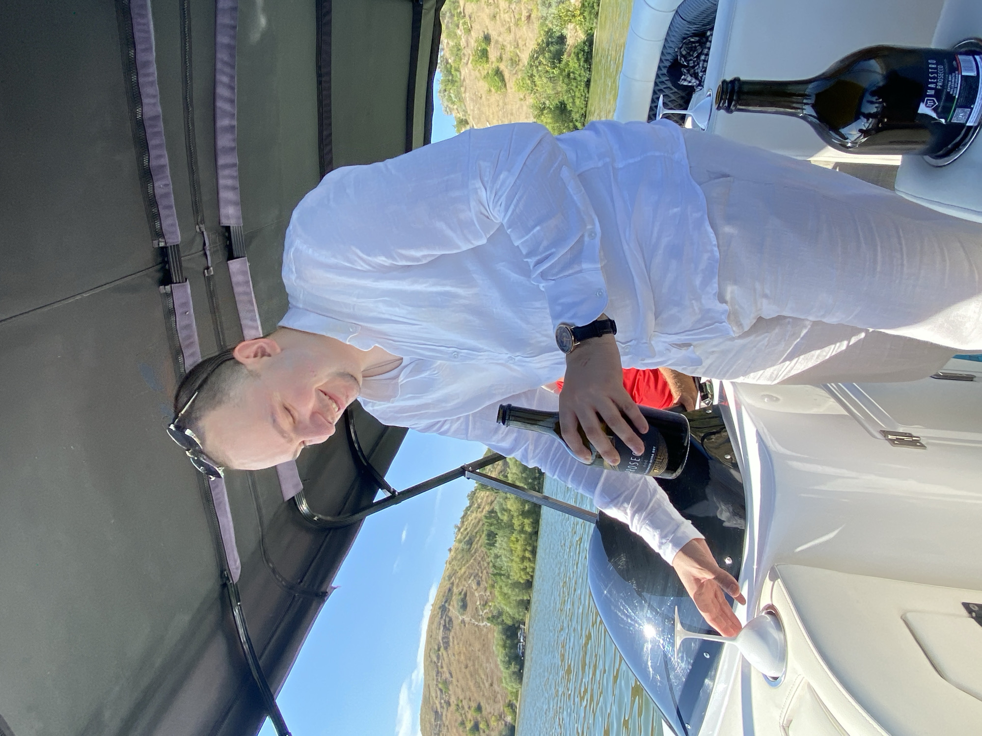 A man in a white shirt and light-colored pants is standing on a boat in Molovata, Moldova. He is holding a bottle of Prosecco and smiling. There is a glass in a cup holder in front of him. The boat is on a body of water with hills in the background. The sky is blue and there are some clouds.