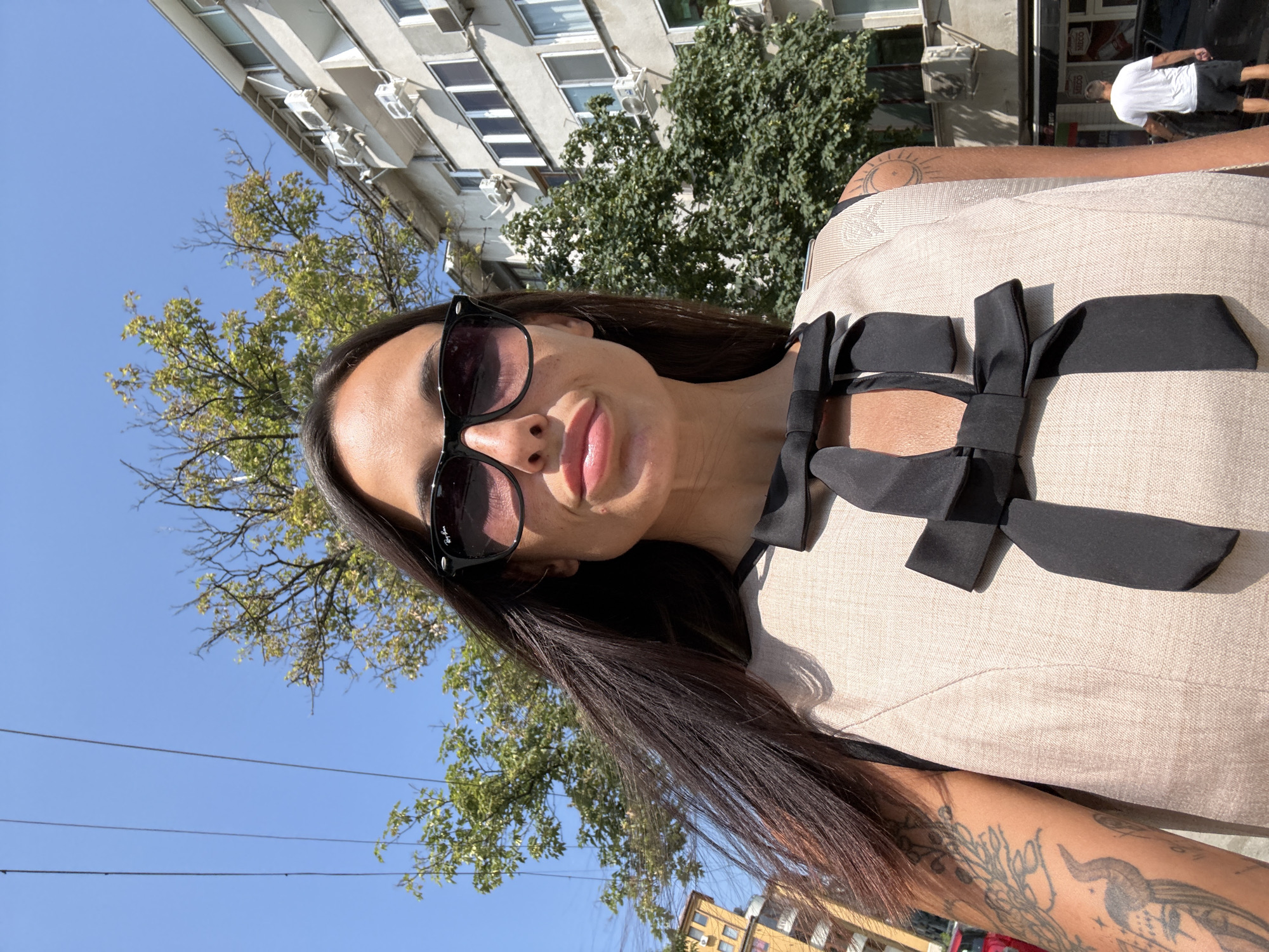 A woman wearing black sunglasses and a light-colored sleeveless dress with black bows is pictured from the chest up. She has long dark hair and visible tattoos on her left arm and shoulder. She appears to be standing outdoors in an urban environment, with buildings and trees in the background. The scene suggests a sunny day. The location is identified as Pleven, Bulgaria.