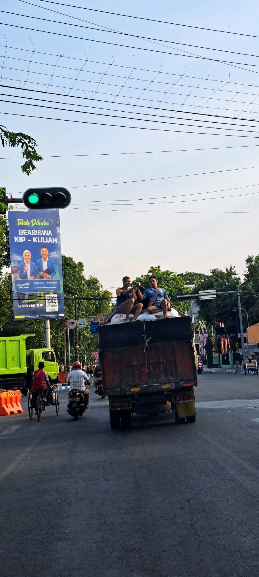 In the city of Pasuruan, Indonesia, on a street under a sky with telephone wires, a traffic light shows green. A billboard announcing "Telah Dibuka BEASISWA KIP - KULIAH" (Scholarships KIP - College is Open) is visible. A large, dark-colored truck is the central subject, carrying several men sitting atop sacks. The truck is in motion, heading away from the viewer. To the left of the truck, a bright green truck is parked, and further left, a man on a bicycle-rickshaw is seen. A motorcycle with a rider wearing a red helmet is also in the lane behind the bicycle-rickshaw. The road surface is asphalt, and the surrounding environment includes trees and some buildings in the background. The scene captures a moment of daily life and transportation in the city.