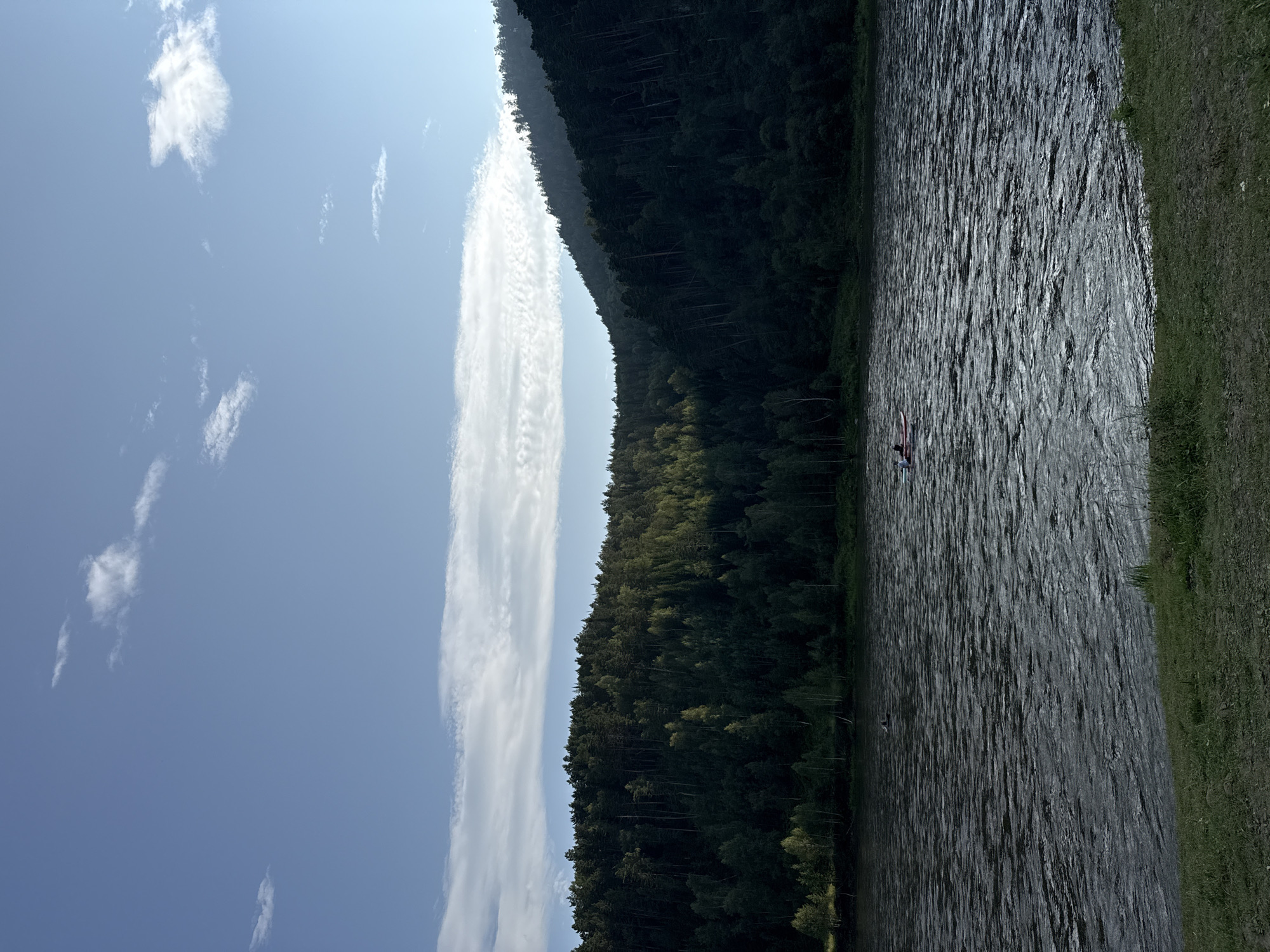 The image depicts a wide, dark river flowing in front of a dense, dark green forest that covers a hillside. The sky is a bright blue with scattered white clouds, and the sun is casting light on a portion of the forest from the left, creating a contrast between the sunlit and shaded areas. In the middle of the river, there is a small red and white boat with two people inside. The foreground shows a grassy bank. The scene is likely captured in Ust-Mana, Russia, given the characteristic Siberian taiga landscape.