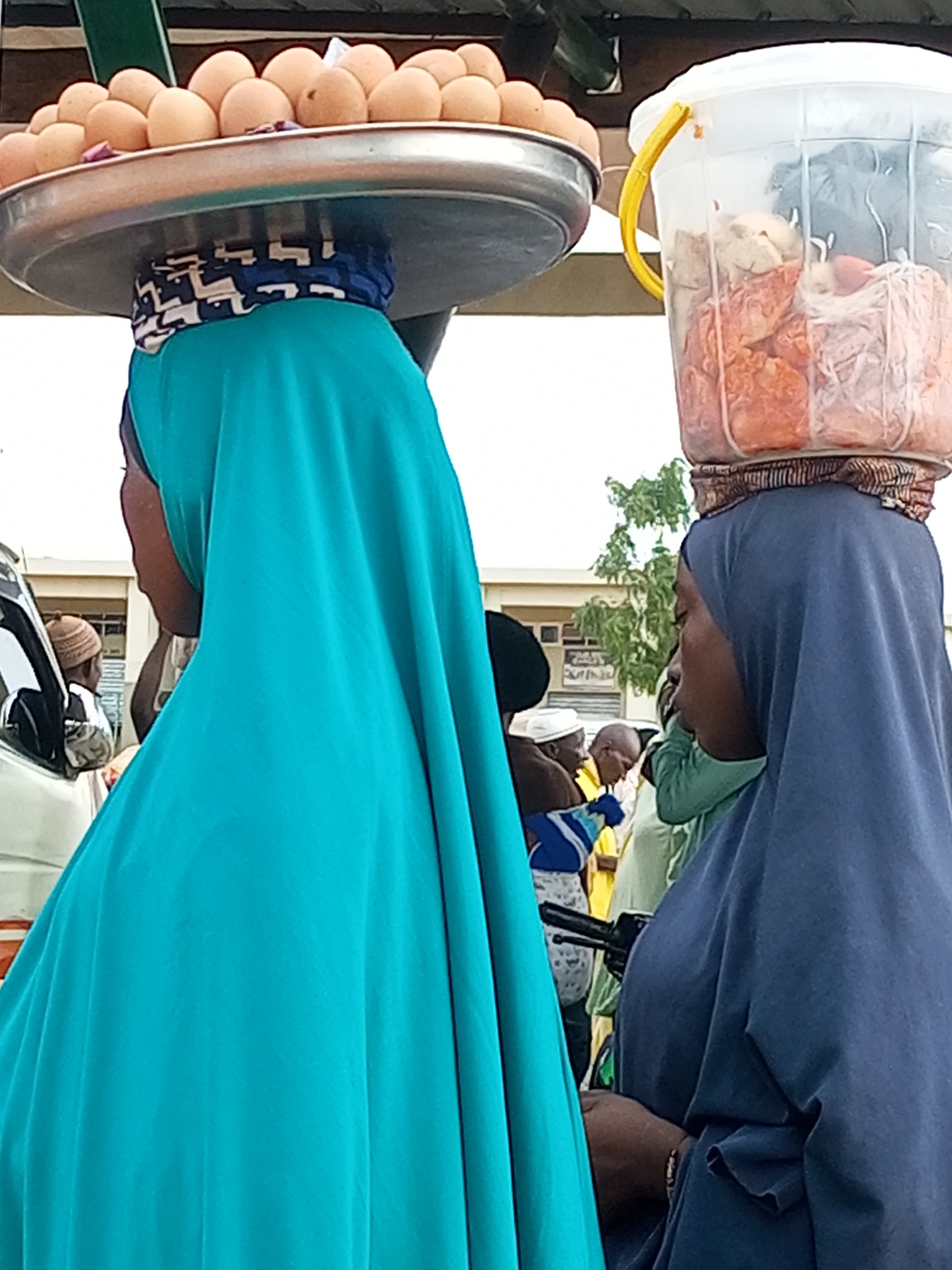 In Gombe, Nigeria, two women are shown wearing hijabs and carrying goods on their heads. The woman on the left has a large metal tray balanced on her head, laden with eggs. She is wearing a turquoise hijab. The woman on the right has a clear plastic bucket filled with what appears to be food items, also balanced on her head. She is wearing a navy blue hijab. They are standing outdoors, with other people and vehicles visible in the background. The scene suggests a market or street-side commerce.