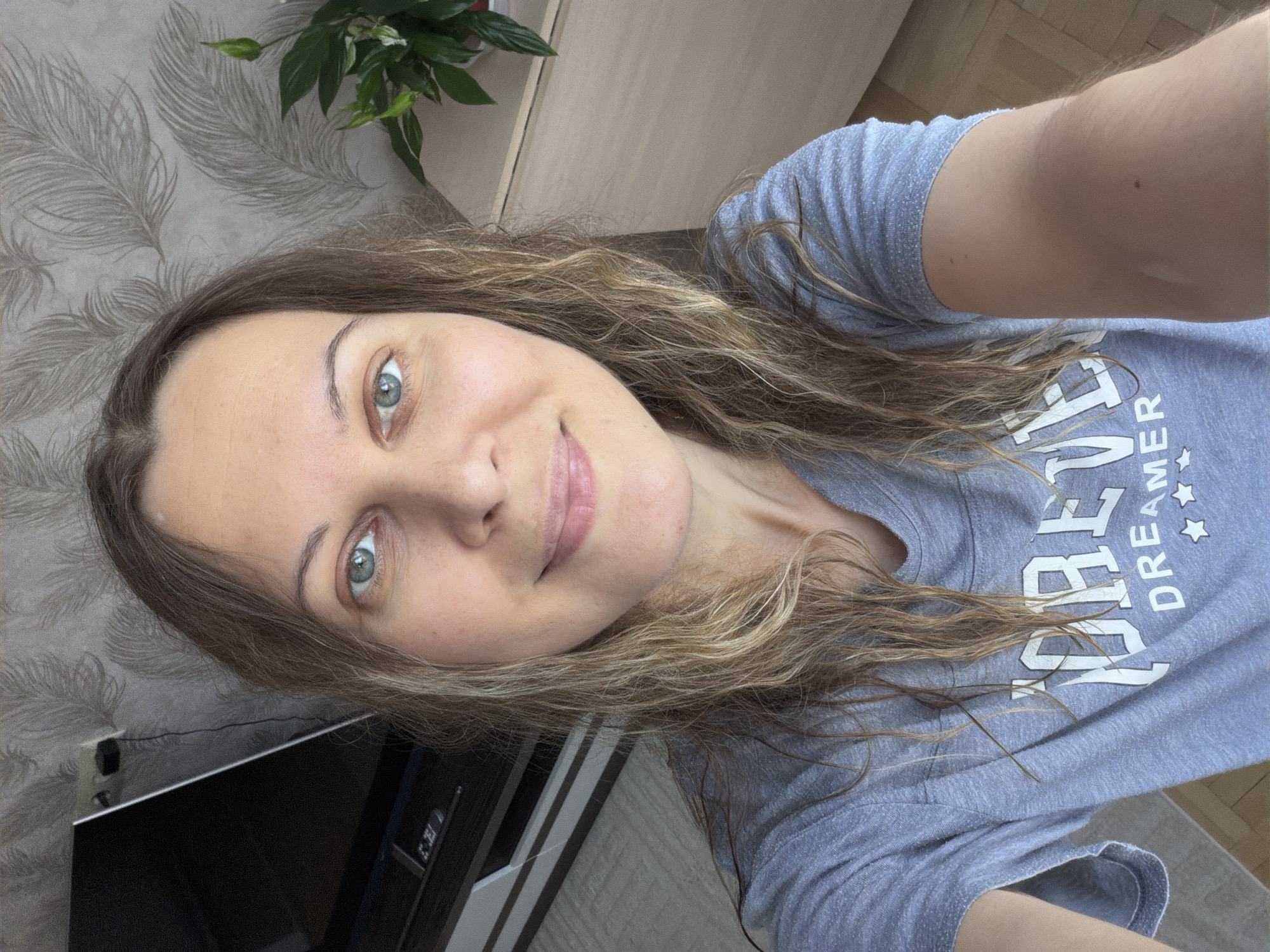A young woman with light brown, wavy hair, wearing a grey t-shirt with white lettering that reads "FOREVER DREAMER", is taking a selfie indoors. She has light blue eyes and is smiling gently at the camera. The background shows a patterned wallpaper, a television set, a wooden cabinet, and a potted plant. The lighting suggests it is daytime. The image is framed as a close-up of the woman's face and upper body. The specific location in the city of Izhevsk, Russia cannot be determined from the visual information provided in the image.