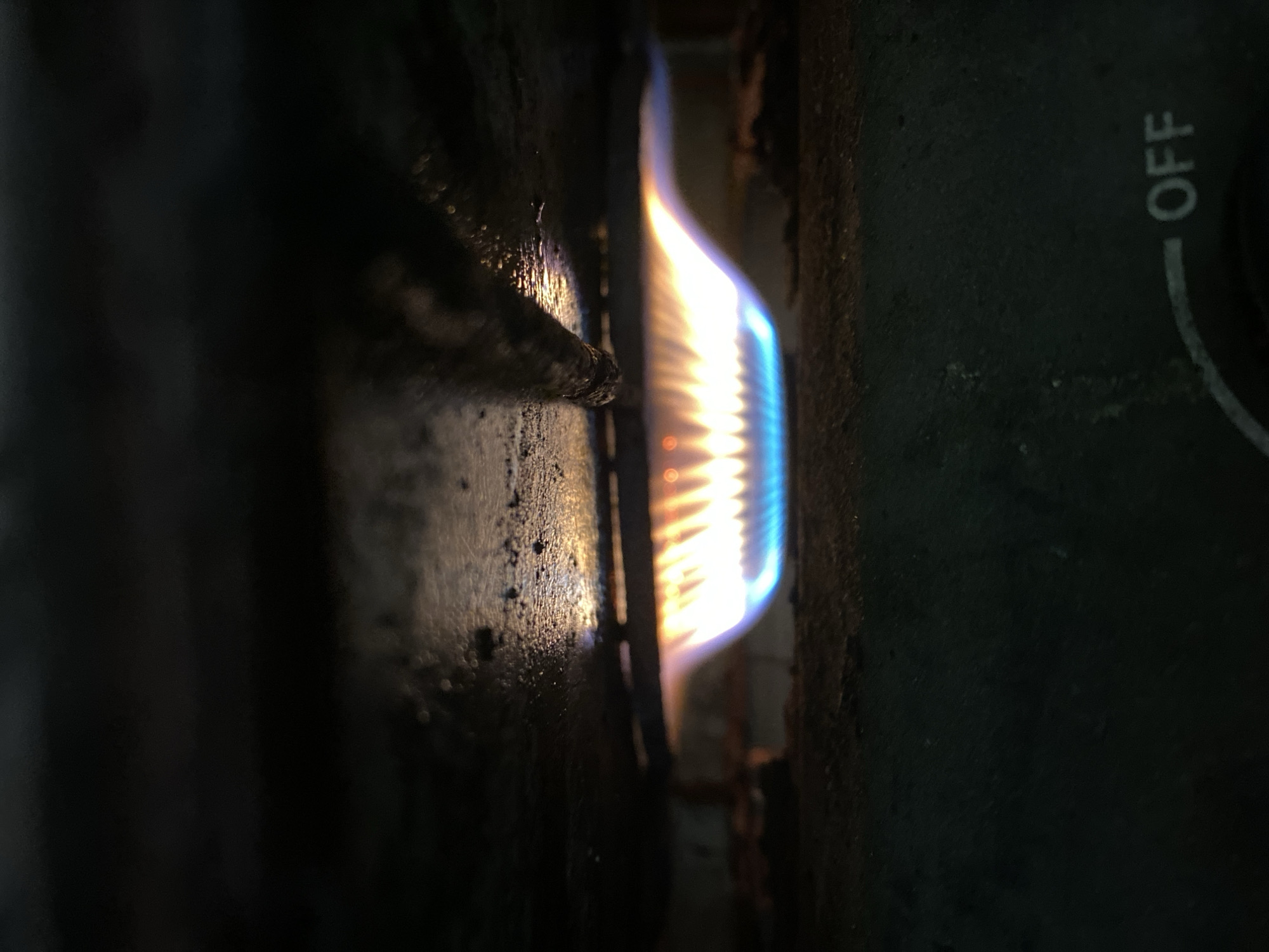 The image shows a close-up, low-angle view of a gas stove burner with a blue and yellow flame. The flame is emanating from a circular burner, producing a curved shape that is lit from below. Above the flame, there is a dark, textured surface that appears to be part of the stove's cooktop, with some light reflecting off it. Below the burner, a dark surface with a dial and the word "OFF" is visible. The background is dark and out of focus, suggesting a dimly lit environment. The image does not contain any information about the city of Kasihan, Indonesia.