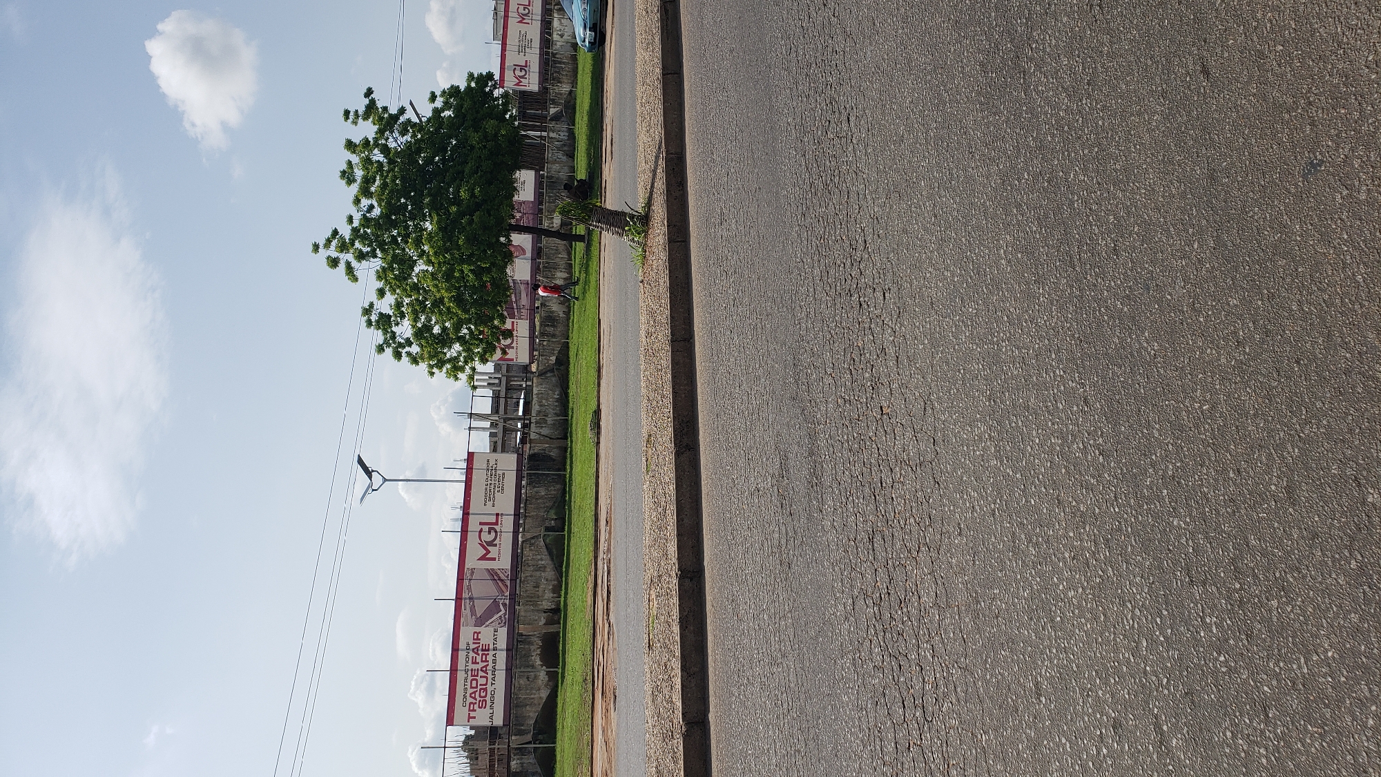 A street view captures a section of Jalingo, Taraba State, Nigeria. In the background, a large banner announces the "CONSTRUCTION OF TRADE FAIR SQUARE, JALINGO, TARABA STATE" and features the logo "MGL". Adjacent to the banner, construction structures are visible. A mature tree with dense green foliage stands prominently in front of the banner and construction site. A single person in a red shirt walks on the grassy area behind a concrete wall and in front of the banner. A light blue vehicle is partially visible on the far right. The foreground consists of a paved road with visible cracks and texture. The sky above is partly cloudy.