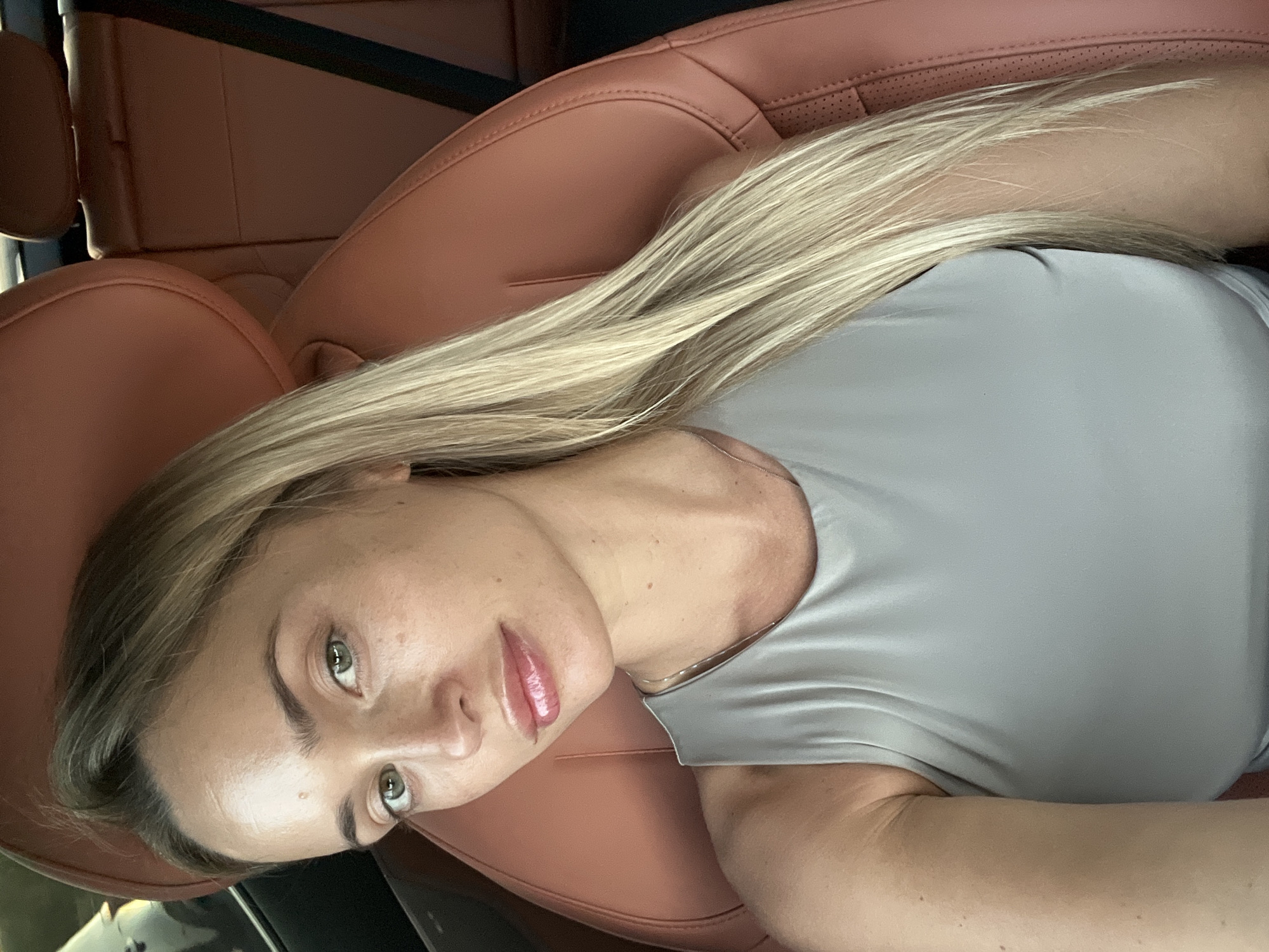 A medium shot image shows a woman with blonde hair and green eyes sitting in the passenger seat of a car. She is wearing a light gray tank top and a silver necklace. The car's interior is visible, with reddish-brown leather seats and a seatbelt. The background suggests an outdoor setting, possibly in Sharm El Sheikh, Egypt, given the bright lighting and what appears to be a coastal or resort area visible through the car window on the left.