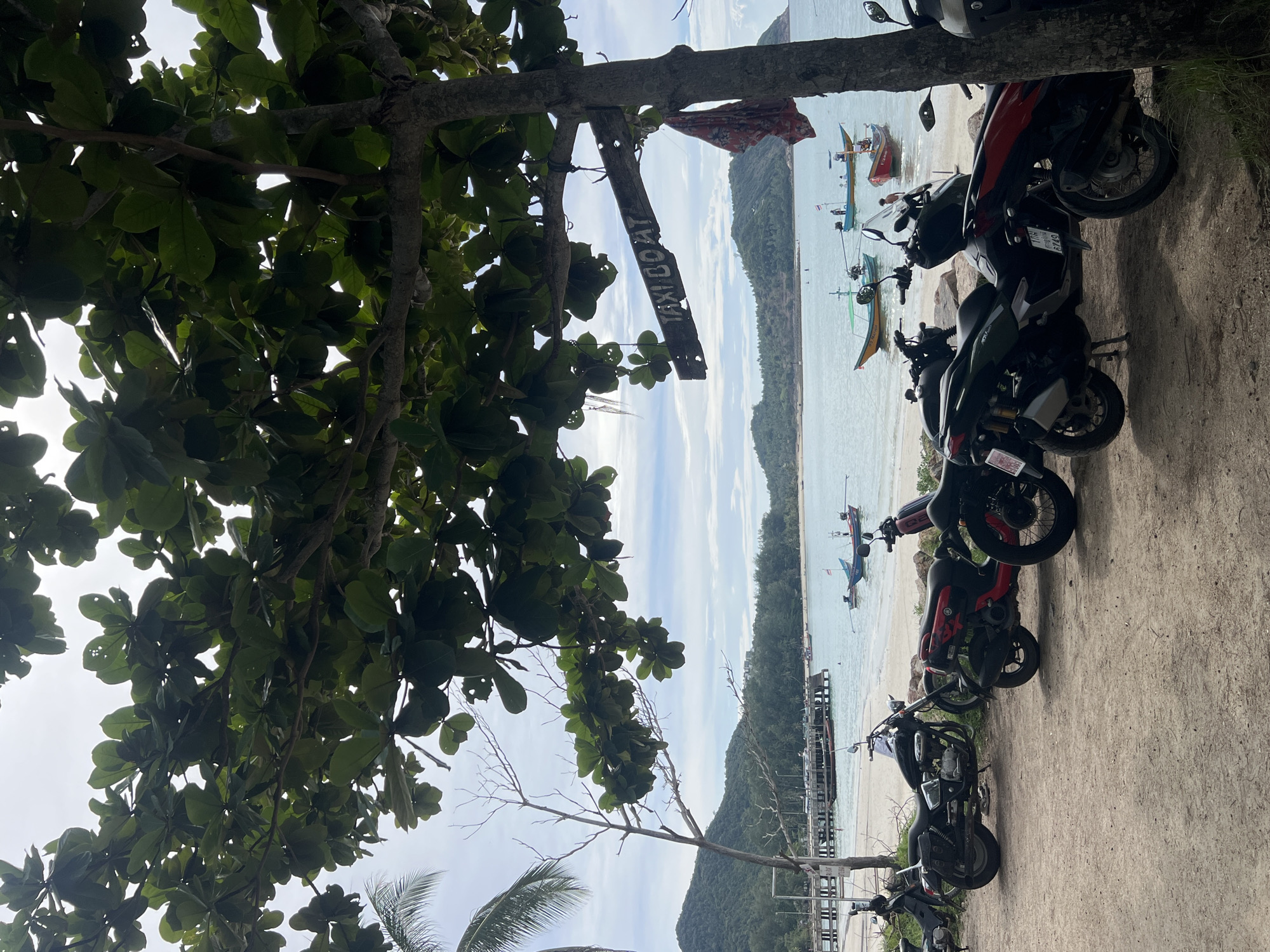 A wooden sign reading "TAXI BOAT" is affixed to a tree trunk in the foreground, to the right of the frame. The sign is weathered and hand-painted. Several motorcycles are parked on a dirt and sand area in the foreground. In the middle ground, a beach meets a calm, turquoise body of water. A wooden pier with a few people on it extends into the water from the left side of the frame. Several small fishing boats are anchored in the water. In the background, lush green hills rise against a cloudy sky. The overall scene depicts a tropical coastal area, likely a transport hub for boat tours.