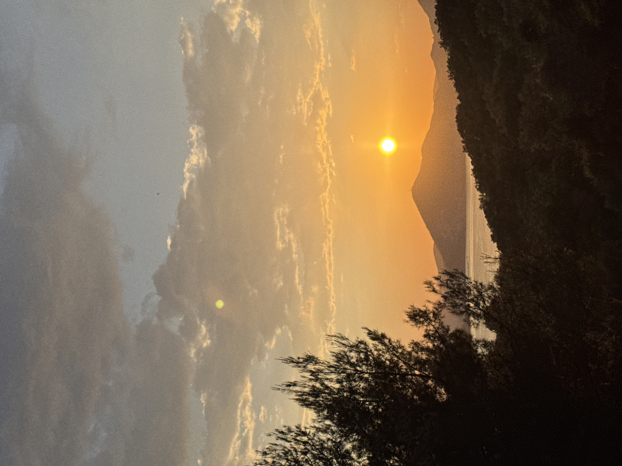 The image depicts a sunset in Sveti Stefan, Montenegro. The sun is positioned near the horizon, between a mountain and the sky, casting an orange glow. Silhouetted trees are in the foreground, and the sea is visible between the mountain and foreground trees. Clouds are scattered throughout the sky.
