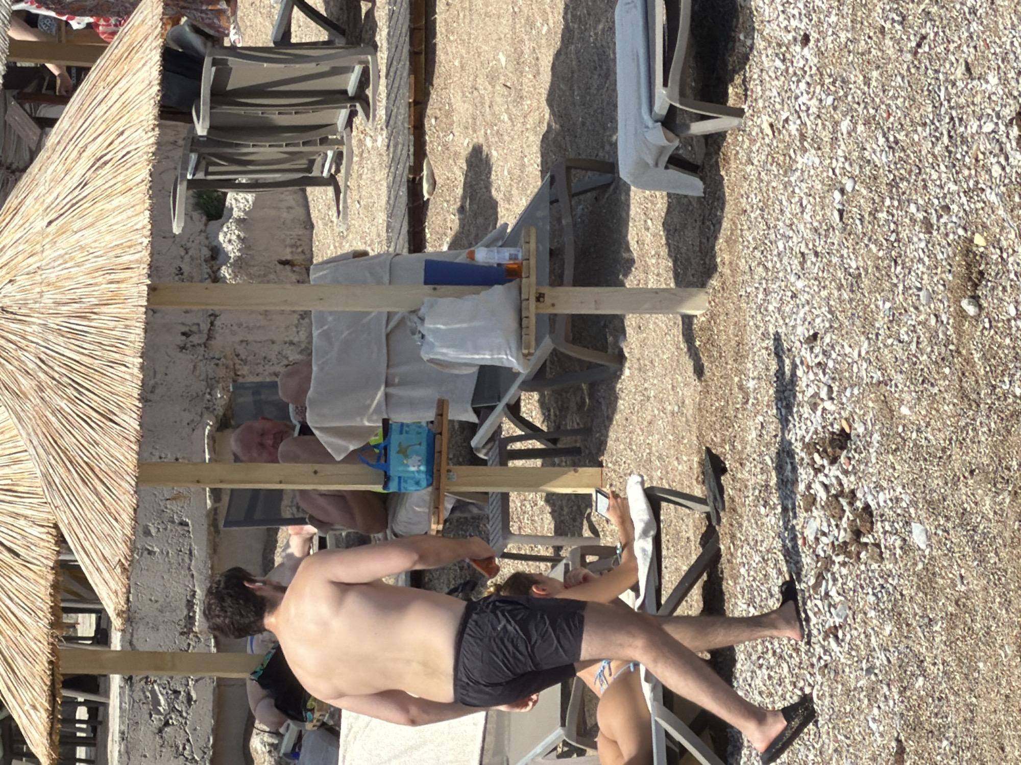 An outdoor scene on a beach in Budva, Montenegro, features several beach umbrellas providing shade. In the foreground, a shirtless man wearing black swim shorts walks away from the camera. To his left, a woman lies on a lounge chair, holding a cell phone. Behind them, another man sits under an umbrella. Beach chairs and a small table with a blue bag and drinks are visible. The ground is covered with small pebbles and sand.