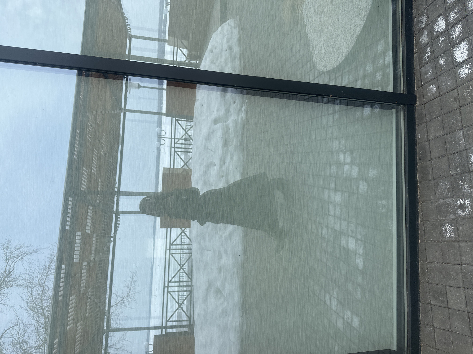 The image displays an exterior perspective of a modern glass-fronted building located in Votkinsk, Russia, likely positioned along the Votkinsk Reservoir. A prominent, dark silhouette of an individual, appearing to wear a long coat or dress with a hood, is reflected on the glass panel, suggesting a person in motion. Through the transparent glass, an outdoor paved patio area is visible, partially covered with patches of snow. This area features decorative dark metal railings with an X-pattern, several light-colored outdoor chairs, and multiple lamp posts with dual curved tops. An overhead pergola-style structure with a slatted canopy is also discernible. Beyond the patio, a vast, flat, snow-covered body of water stretches towards the horizon under an overcast sky. Bare tree branches are visible against the sky on the left. The ground directly in front of the glass consists of dark gray rectangular pavers, exhibiting scattered white residue.