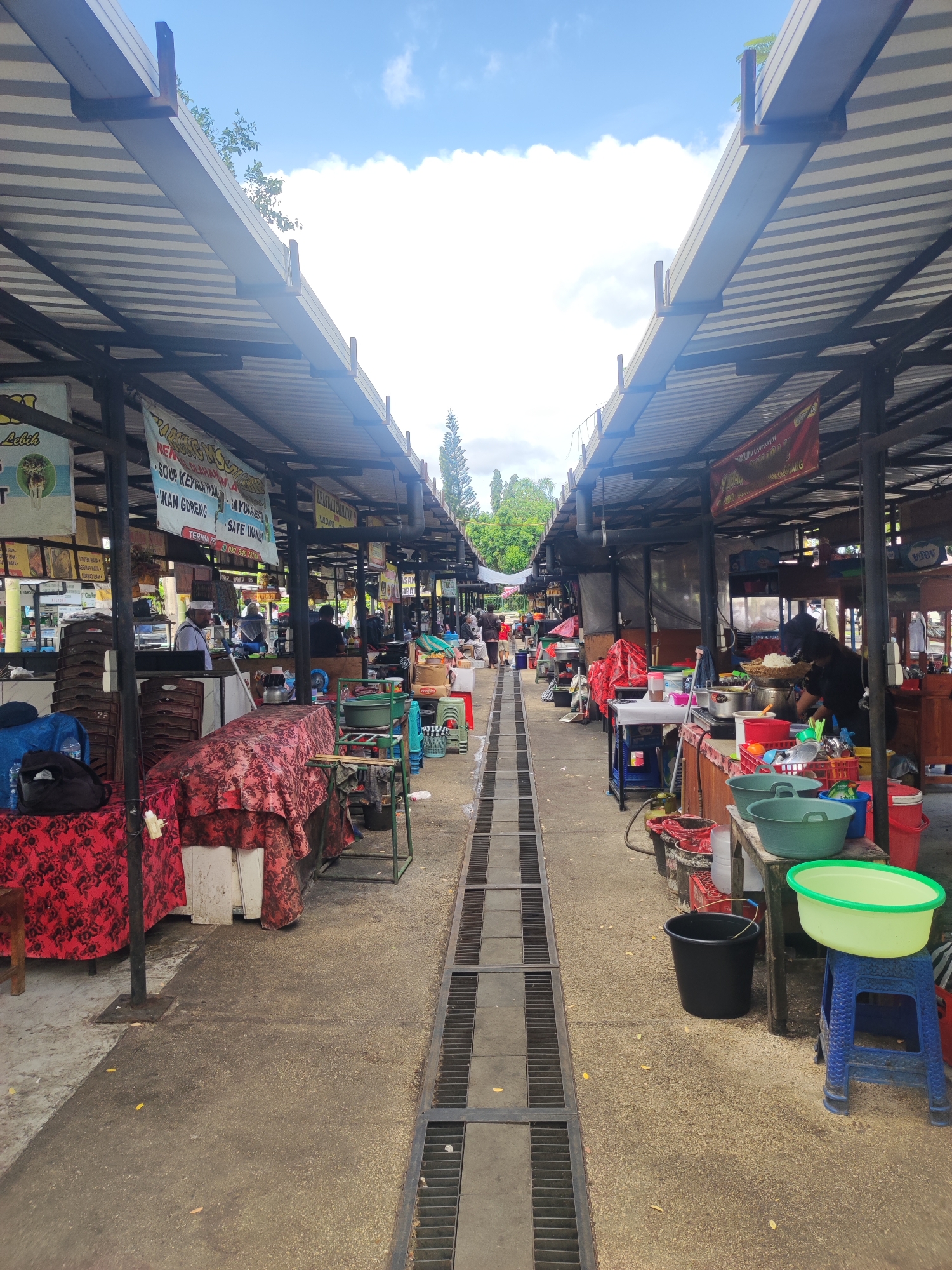 The image depicts an outdoor market in Gianyar, Indonesia, with stalls lining both sides of a central walkway. A drainage system runs down the center. The stalls are covered by corrugated metal roofs supported by metal frames. Various vendors are present, some preparing food, others tending to their goods. Tables covered with cloths, stacks of chairs, cooking equipment, buckets, and basins are visible at the stalls. Banners with Indonesian writing are hung at some stalls. The sky is partially cloudy.