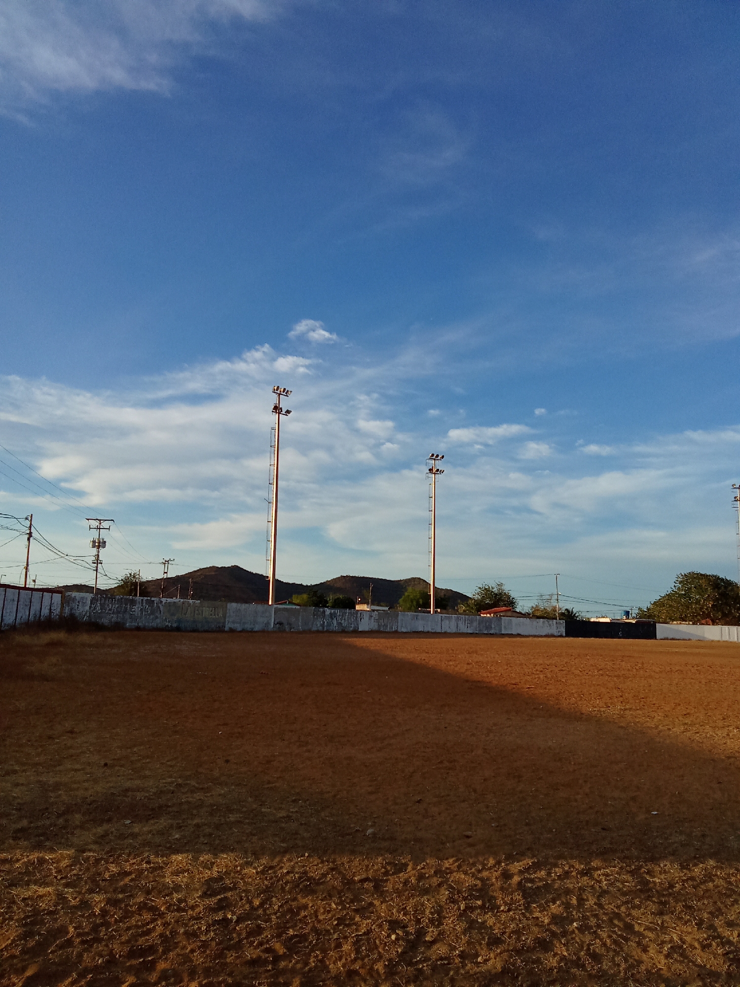 A daytime shot of a mostly barren, reddish-brown field, likely a sports field, in Juan Griego, Venezuela. A low, whitewashed wall runs along the periphery of the field, behind which are power lines and utility poles. Beyond the wall, a low mountain range is visible. The sky is blue with scattered white clouds. Three tall metal light posts with multiple lights are positioned along the perimeter of the field. The ground cover appears sparse and dry.