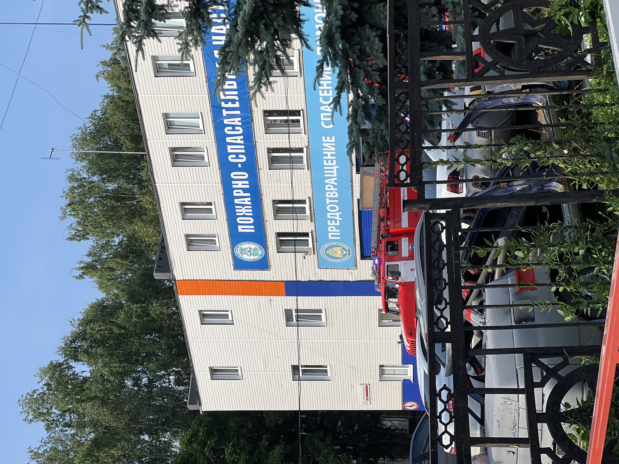 The image depicts the exterior of a building in Izhevsk, Russia, likely a fire and rescue station given the presence of a red fire truck and banners with Russian text that translates to "FIRE AND RESCUE" and "PREVENTION RESCUE." The building is a multi-story structure with light-colored siding and orange and blue accents. In front of the building, several damaged or wrecked cars are visible behind a black metal fence. The cars appear to be in a state of disrepair, with some having open doors and visible damage. A fire truck, with flashing lights, is parked in front of the building. The environment is outdoors, with trees and a clear blue sky visible in the background. The scene suggests a recent incident or an area where damaged vehicles are stored.