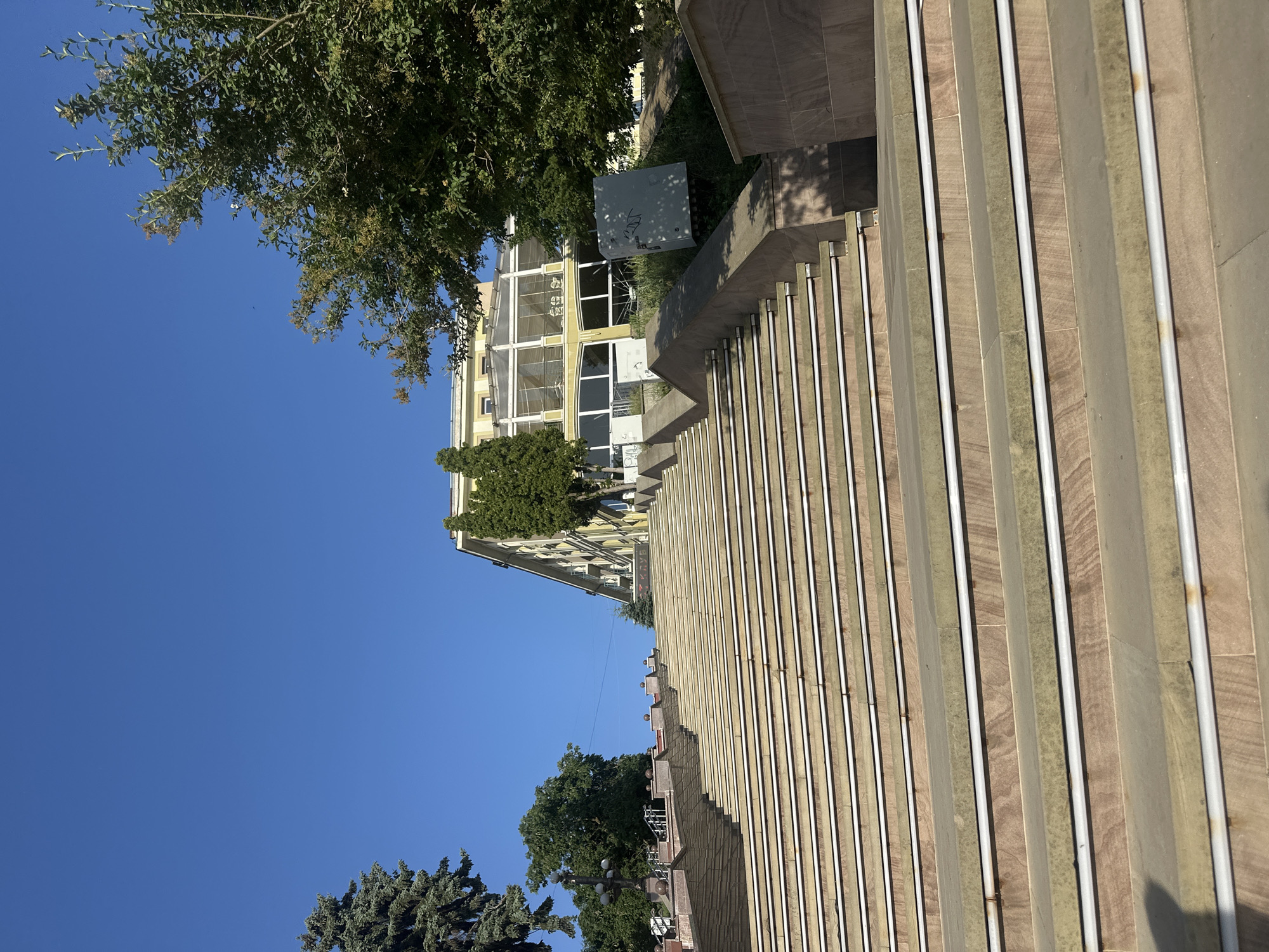 The image depicts an outdoor scene with a clear blue sky. In the foreground, a set of stone stairs with metal railings runs diagonally across the frame. The stone appears to be light brown and grey, with a textured surface. The metal railings are silver and run parallel to the edge of each step.
Behind the stairs, a building with a yellowish facade is visible. It has multiple windows with dark frames and a light-colored trim. Trees with green foliage are interspersed between the stairs and the building, as well as in the background. One tree in the foreground on the left is quite large and leafy. A gray utility box with some markings is situated on the ground near the building, partially obscured by greenery.
Further in the background, more trees and parts of other buildings can be seen. The overall impression is of a sunny day, likely in the late morning or afternoon, given the shadows cast by the trees and the lighting on the buildings. The scene appears to be in a public space, possibly a park or an area around a building in Ternopil, Ukraine. There is no apparent activity or event taking place, and the image seems to be a static view of the environment. No specific emotions are conveyed, and no readable text is clearly visible.