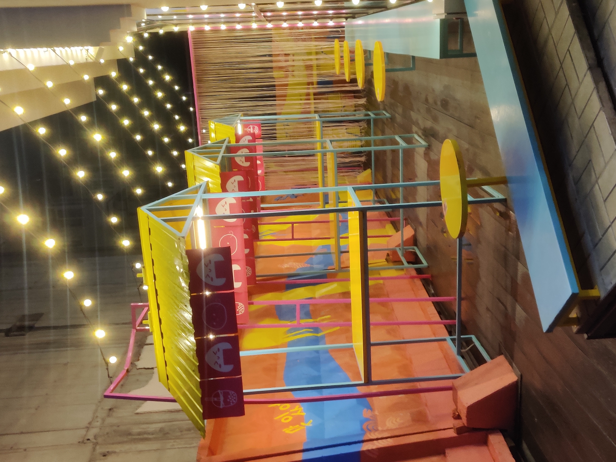 The image depicts an outdoor food court with a whimsical and colorful design, likely situated in a contemporary urban setting. The food stalls are constructed with bright yellow roofs and light blue and pink metal frames, resembling small, stylized houses. Each stall features a counter space and is adorned with red banners displaying cartoonish images of onigiri (rice balls) and bubble tea.
The background reveals a textured concrete wall on the left and a curtain of thin, light-colored strings on the right, creating a softer, atmospheric backdrop. Numerous strings of warm-toned fairy lights are strung across the scene, illuminating the area and contributing to a festive ambiance, especially given the twilight or evening lighting.
In the foreground, there are several small, round yellow tables and long, light blue benches with yellow trim, suggesting seating arrangements for patrons. The ground is made of dark wooden planks, which appear slightly damp, possibly from recent rain or cleaning. A large, orange structure with blue and yellow abstract painted designs is visible on the far left, possibly a shipping container repurposed as part of the food court's structure.
There are no people visible in the image. The overall impression is that of a trendy, perhaps newly established, outdoor dining or entertainment space, designed to be visually appealing and engaging. The specific location in Yekaterinburg, Russia, is not visually indicated by any landmarks or text within the image itself.