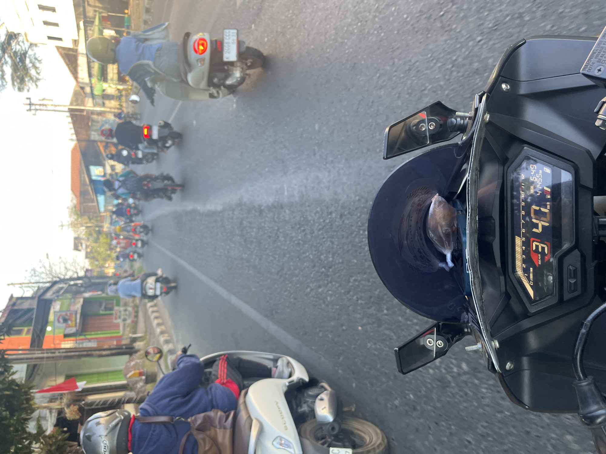 The image captures a POV shot from a motorcycle dashboard, showing a street scene in Pecangaan Wetan, Indonesia, during daylight. The dashboard displays the current gear as '3' and speed as '40 km/h', with a total time of '6:49' and distance of '41455 km'. Multiple motorcycles with riders wearing helmets are traveling on a paved road, suggesting a busy traffic environment. Buildings line the side of the road, some appearing commercial with signs and awnings, and others residential. A red and white flag is visible on a building to the left. The road has painted lane markers and a textured surface. The sky is bright, indicating clear weather.