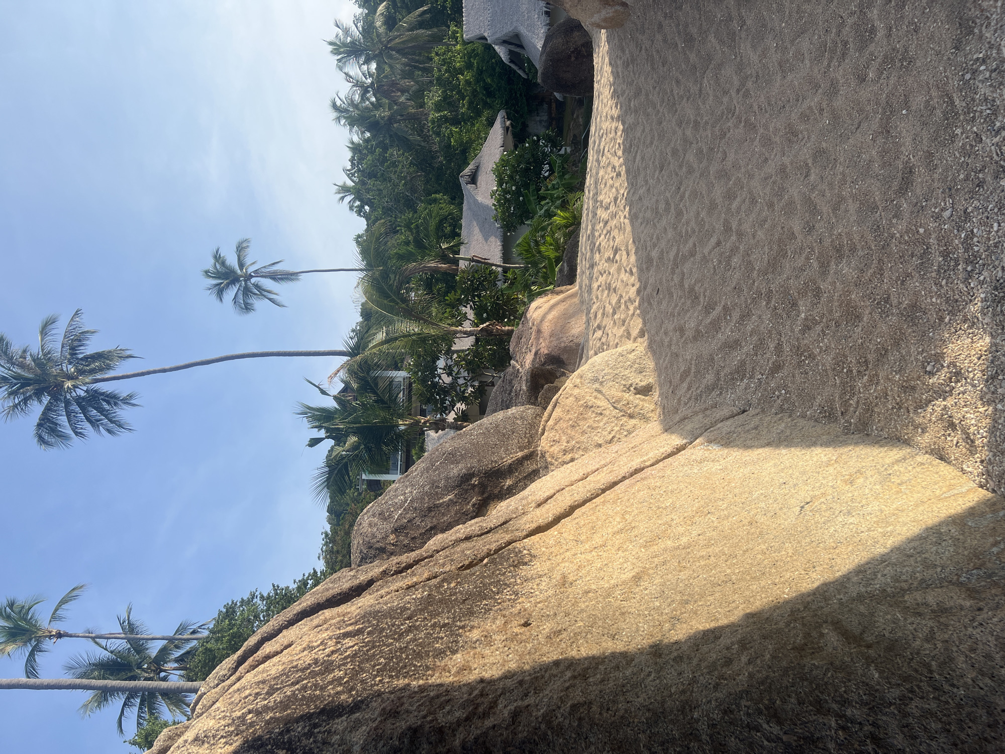 The image depicts a tropical beach scene in the Ko Samui District of Thailand, characterized by large granite boulders, fine sand, and lush vegetation. In the foreground, a prominent granite boulder dominates the left side of the frame, casting a long shadow across the sandy path leading into the scene. The path itself is composed of fine, light-colored sand with visible ripple marks from the wind or water.
Beyond the boulder and sand, several thatched-roof bungalows are visible, nestled amongst dense green foliage and tall palm trees. These structures suggest a resort or residential area. The palm trees, characteristic of tropical environments, are scattered throughout the landscape, with their fronds reaching towards a clear blue sky. The presence of these bungalows and well-maintained vegetation indicates human habitation and development within this natural setting. The overall environment is serene and sunlit, suggesting a warm, tropical climate. There are no people or animals visible in the frame.