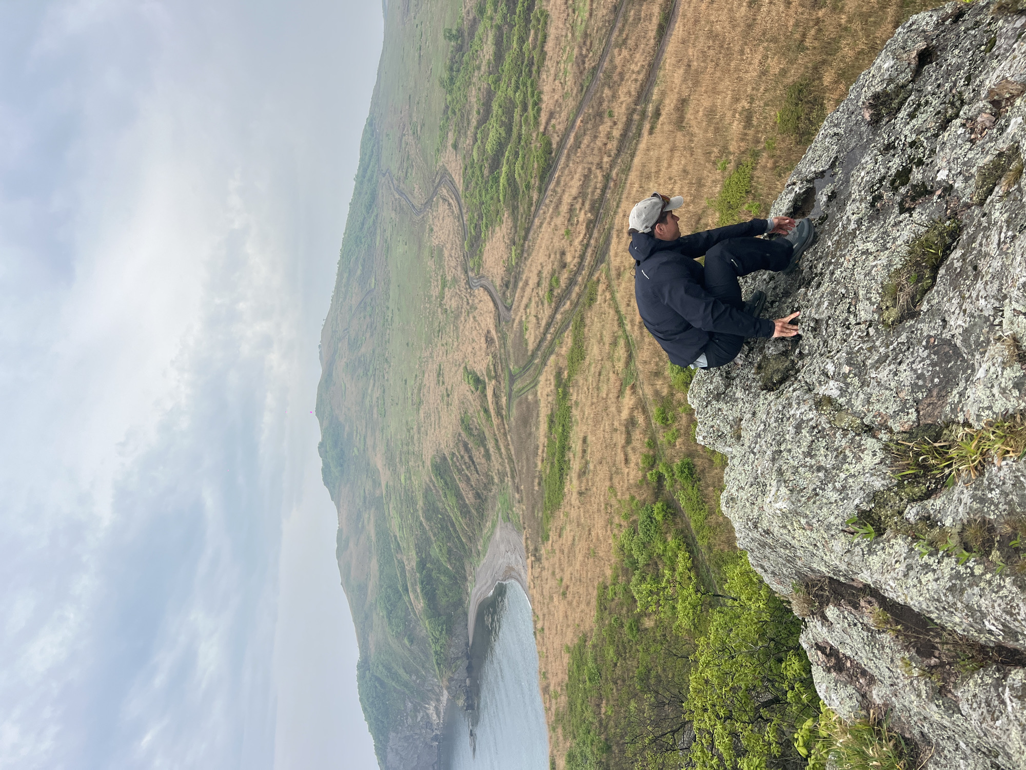 The image shows a man sitting on a rocky cliff overlooking a landscape. The man is wearing a dark jacket, dark pants, a light-colored baseball cap, and dark shoes. He is sitting cross-legged and looking out at the view. The cliff is made of grey rock and is covered in some moss and small plants.
In the background, there is a hillside with a mixture of green and brown vegetation. A winding dirt track can be seen traversing the hillside. At the bottom of the hill is a small beach and the ocean, which stretches out to the horizon. The sky is overcast.
The location is identified as being in the city of Gvozdevo, Russia.