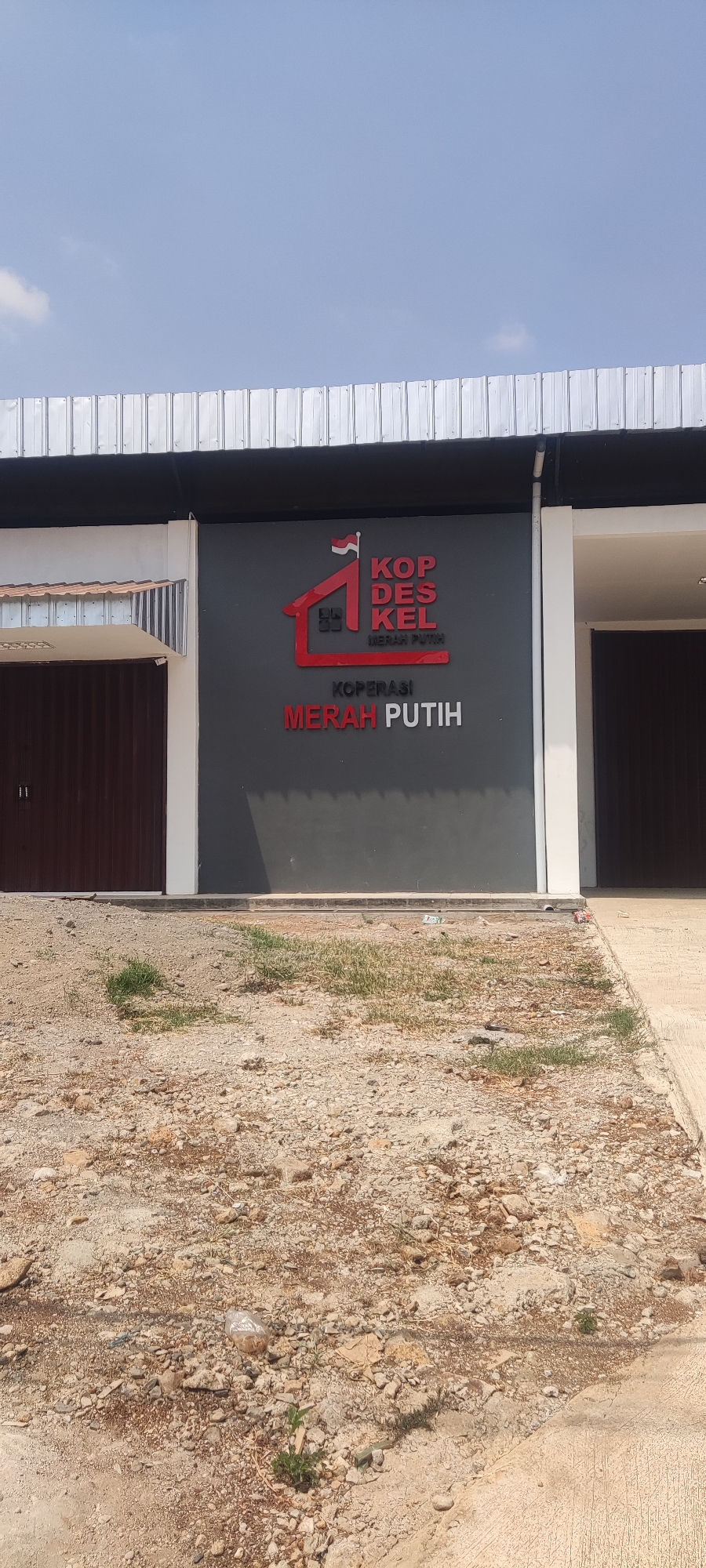 The image shows a building with a dark gray facade featuring a prominent red and white logo and text. The logo is a stylized red house with a flag and the text "KOPDESKEL" and "MERAH PUTIH" in red lettering. Below this, in white lettering, is "KOPERASI MERAH PUTIH". To the left of the facade is a dark brown roll-up door. The building has a white trim and a metal roof with visible support structures. The ground in front of the building is unpaved, with dirt, rocks, and some patches of dry grass. A concrete path is visible to the right of the building. The sky above is clear blue with a few wispy clouds. This scene is located in Panongan Lor, Indonesia.