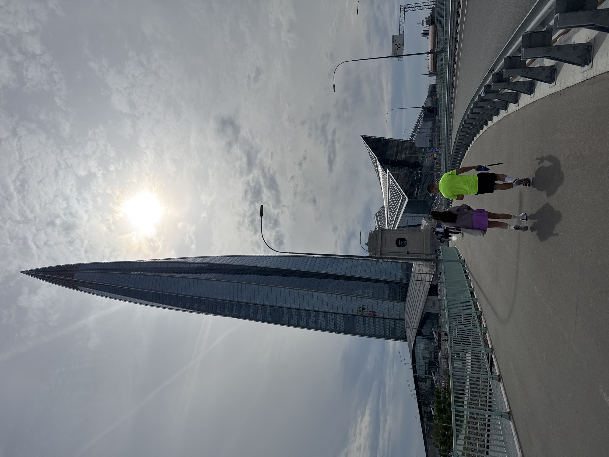 A wide-angle, rotated long shot depicts a sunny scene in Saint Petersburg, Russia, focusing on the Lakhta Center skyscraper and a pedestrian walkway. The Lakhta Center, a modern glass skyscraper with a sharp, pointed top, dominates the left side of the frame. The sky is filled with clouds, and the sun is shining brightly, creating a glare.
On the right side of the frame, a pedestrian walkway is visible. Two people are walking on the walkway. The person closest to the foreground wears a neon green shirt and dark shorts and carries a small object, possibly a dog leash. The other person wears a lavender-colored top and skirt. Shadows of both people are visible on the path. A green metal railing borders the walkway. A highway runs alongside the walkway.
