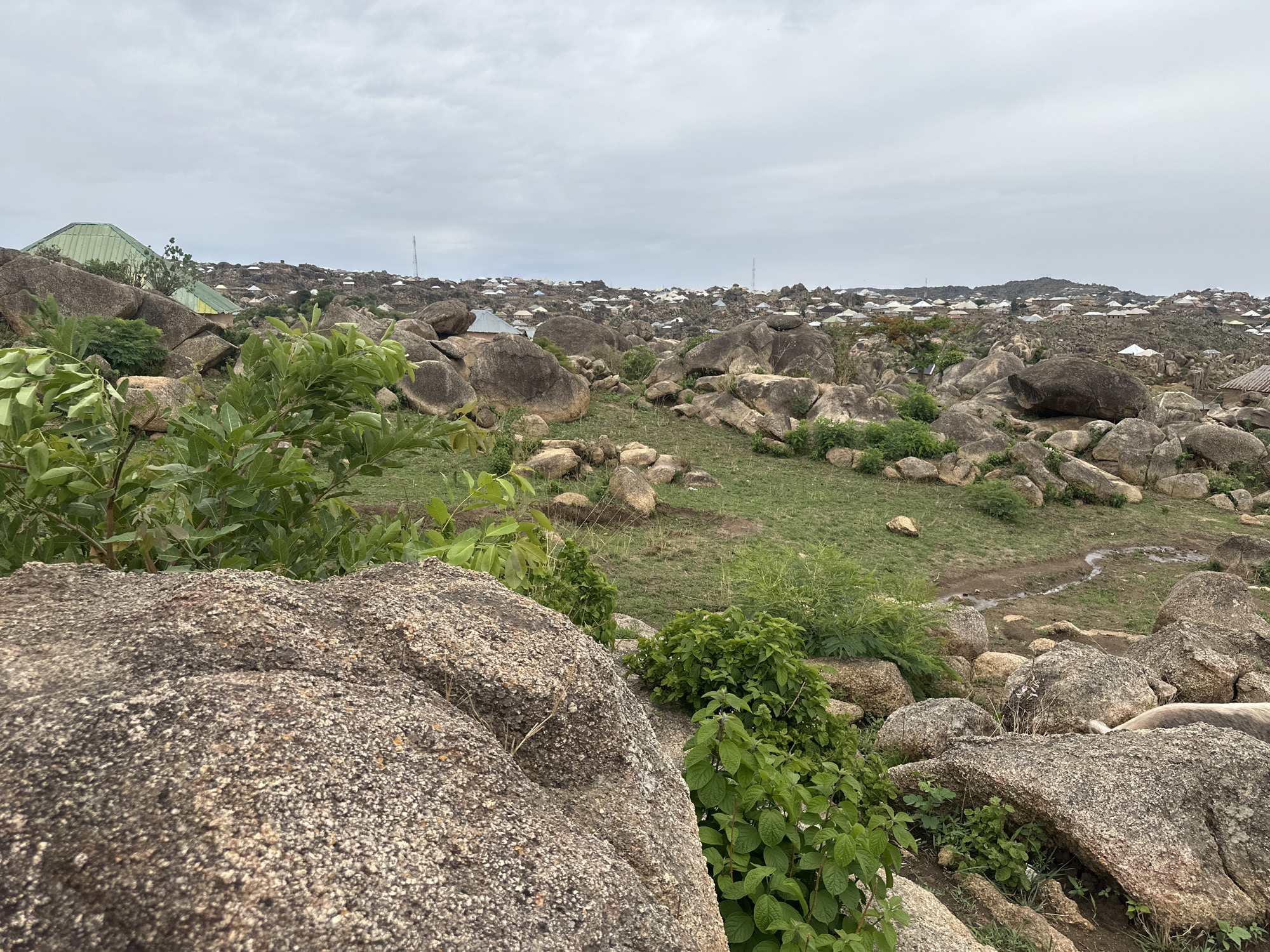 The image shows a landscape in Jos, Nigeria, characterized by large, rounded granite boulders interspersed with patches of green vegetation. In the distance, a dense settlement of low-rise buildings with various roof colors is visible under a cloudy sky. Some buildings have green roofing. There are multiple cell towers visible in the distance. The foreground consists mainly of the weathered surfaces of the rocks, some with scattered green foliage. A small, muddy stream or drainage channel cuts through the green space between the rocks.