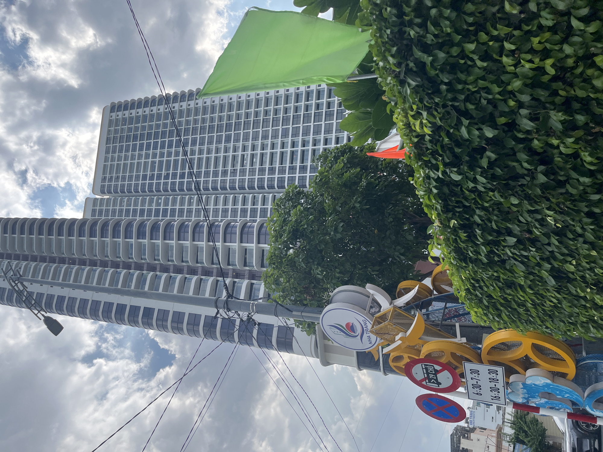 In Nha Trang, Vietnam, a modern cityscape is depicted under a cloudy sky. Two tall, white high-rise buildings dominate the background, their windows forming a grid-like pattern. In the foreground, a lamppost supports a network of power lines. Attached to the lamppost is a circular sign with the text "NHA TRANG" and a stylized logo featuring a sun setting over water. Below this, a collection of decorative metal sculptures in yellow and blue hues is visible, along with traffic signs indicating parking restrictions and bus times. A lush green hedge lines the bottom of the image, partially obscuring a dark-colored car on the left. A bright green flag stands tall on the right, and a smaller orange flag can be seen peeking out from behind the hedge. The overall impression is one of urban development juxtaposed with natural elements and local cultural markers.