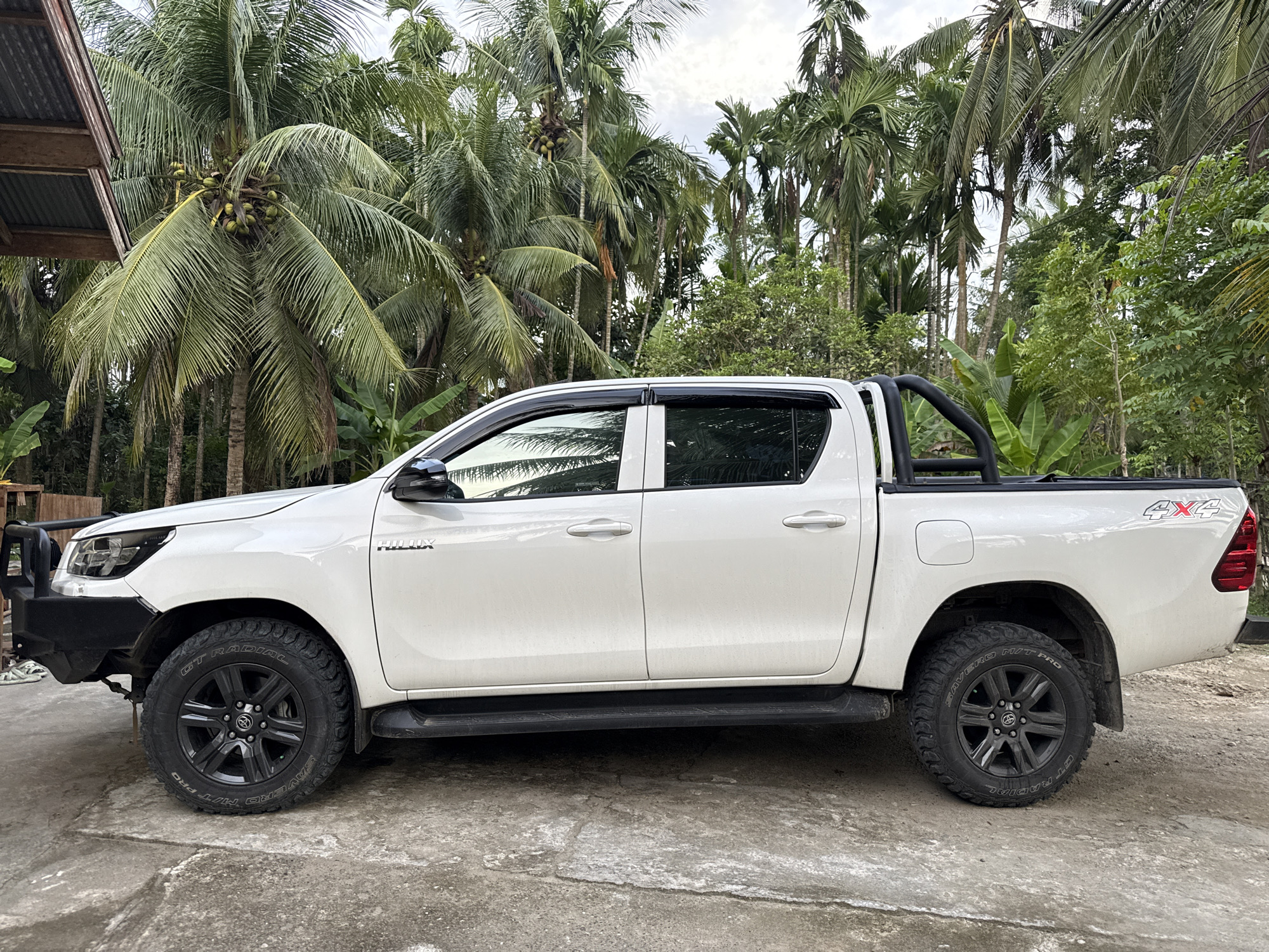 A white Toyota Hilux pickup truck is parked on a concrete surface. The truck has black wheels and a black bull bar on the front. It also features a black sports bar in the bed. The words "HILUX" and "4x4" are visible on the vehicle. In the background, there is a dense array of palm trees and other tropical vegetation. A partial structure with a corrugated roof is visible to the left. The location is identified as Pulorungkom, Indonesia.