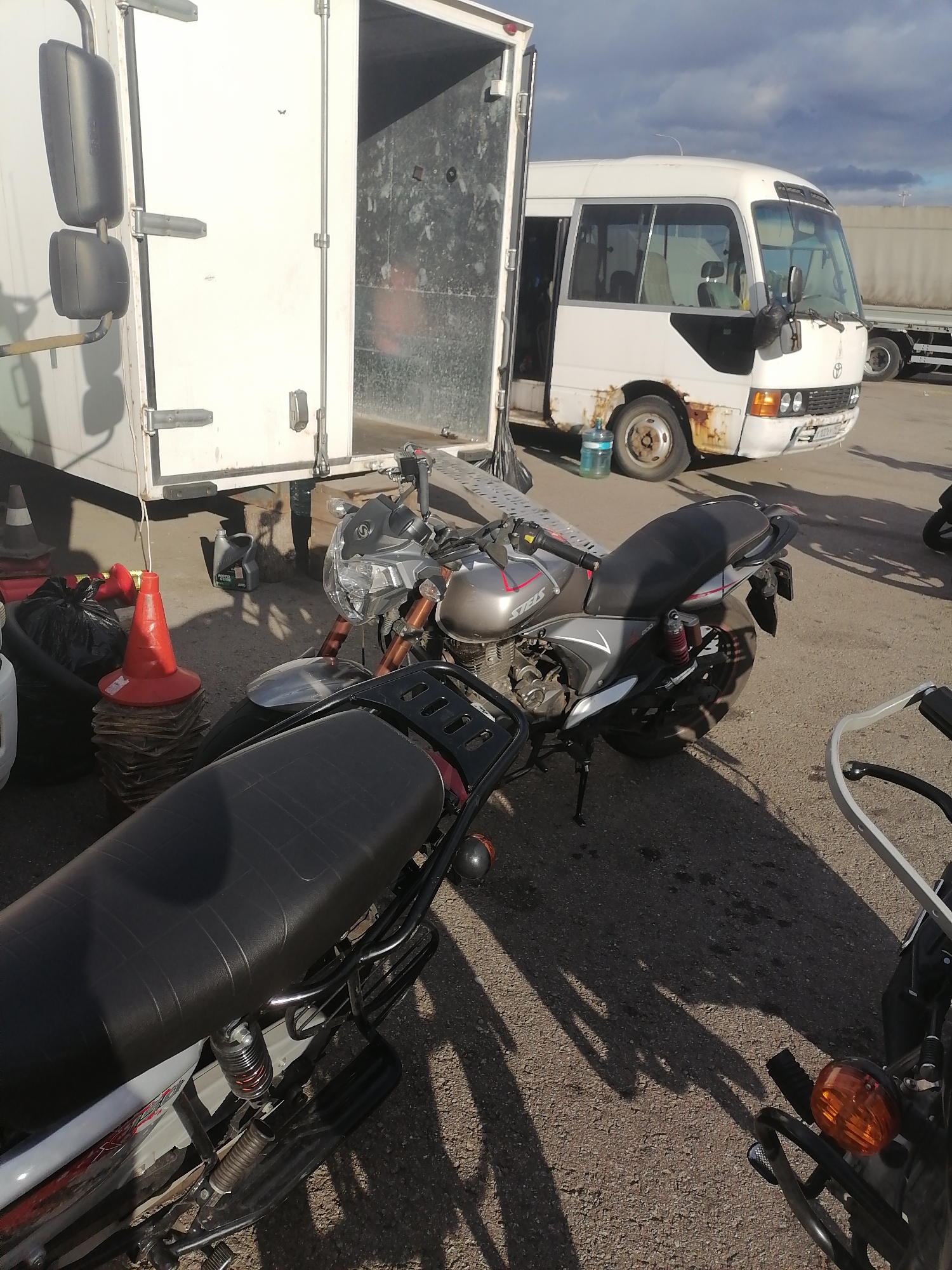 The image shows a street scene in Moscow, Russia, under a partly cloudy sky. In the foreground, there are two motorcycles parked on asphalt. The motorcycle closer to the viewer has a black seat and a silver and red body. Behind it, another motorcycle is visible, primarily silver with black accents. To the left, a white truck with its rear door open is parked. Further in the background, a white minibus with signs of rust on its body is parked. Orange traffic cones and black garbage bags are situated near the truck, and a bottle of water is on the ground next to the minibus's front wheel. The scene suggests a loading or unloading area, possibly related to transportation or event setup.