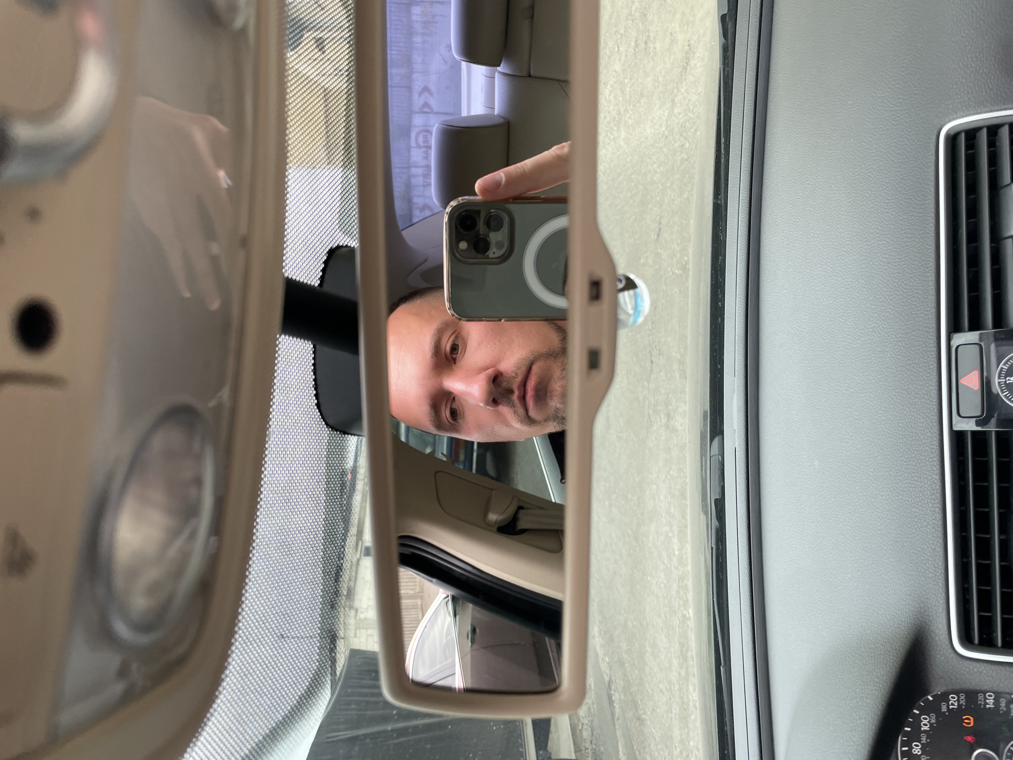 A man is taking a selfie in the rearview mirror of a car. The man is in the driver's seat, and the car appears to be parked. In the background of the mirror, there are other cars and buildings, indicating an urban setting. The car interior is visible in the foreground, including the dashboard and a part of the steering wheel. The man is holding a smartphone, which is reflecting the car's interior and his face. The image appears to have been taken during the daytime, with overcast or cloudy skies.