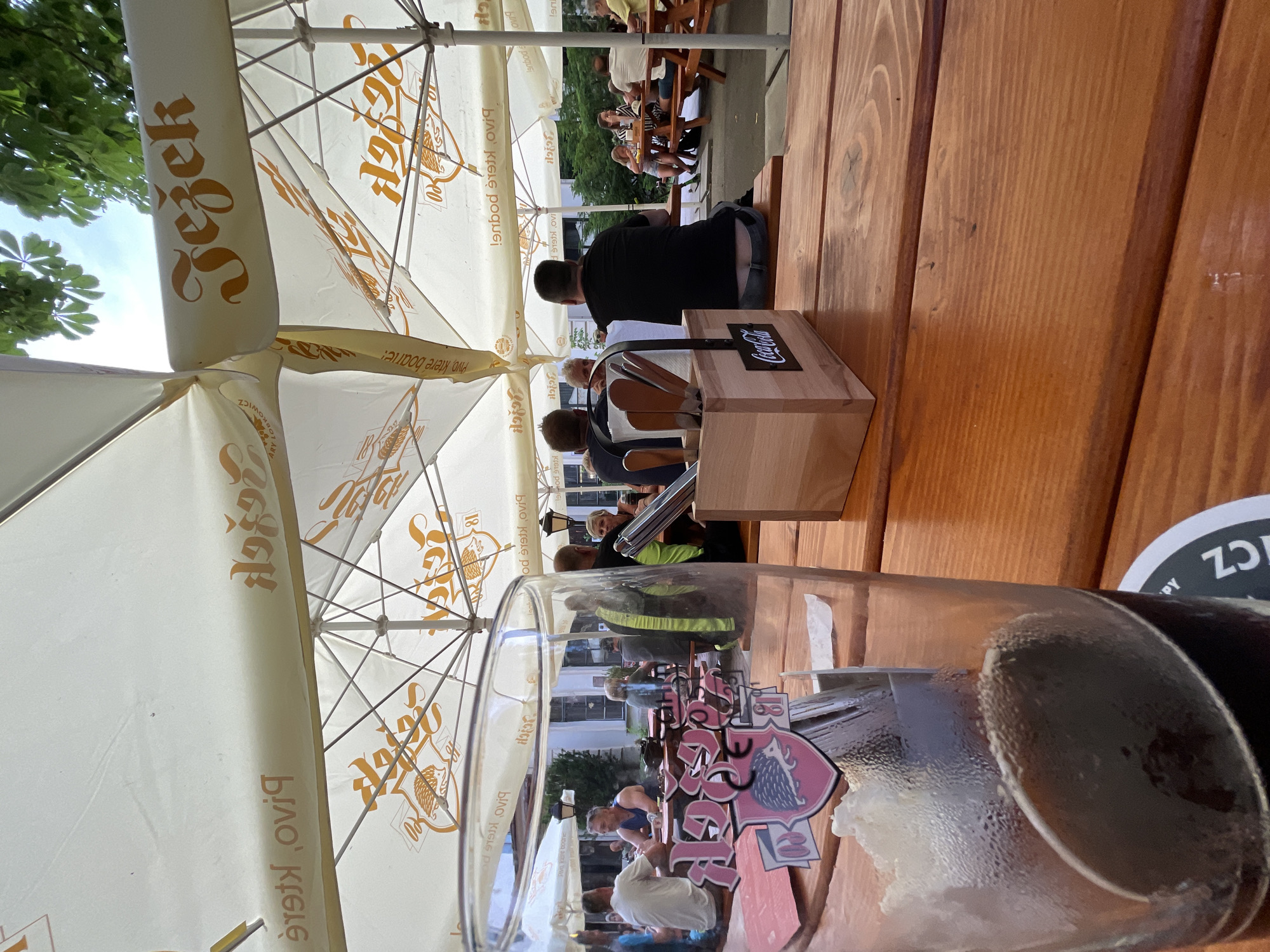 The image depicts an outdoor seating area of a beer garden, likely located in Jihlava, Czechia, under large white umbrellas that bear branding. In the foreground, a glass of dark beer with foam sits on a wooden table, partially obscuring a colorful sticker on the glass with a hedgehog logo and the text "Jezek 18". A wooden holder containing cutlery and a Coca-Cola logo is also on the table. Several patrons are seated at picnic tables in the background, enjoying drinks and socializing. The environment suggests a relaxed, casual atmosphere, possibly during daytime or early evening.