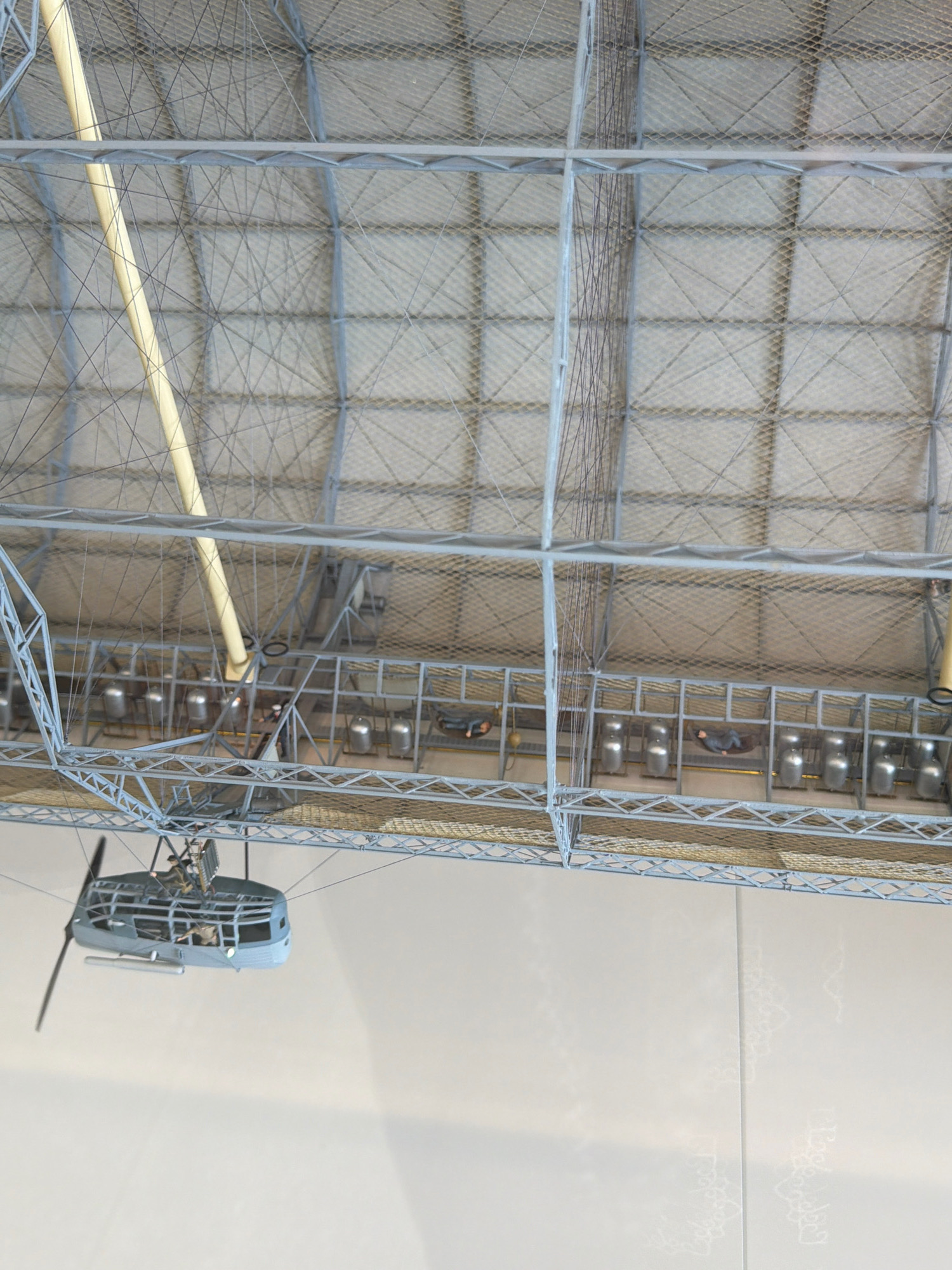 This is an interior view of a hangar, likely the Zeppelin Museum in Friedrichshafen, Germany, showcasing a detailed model of a historic airship hangar. The scene is viewed from a high angle, looking down at the structure.
The hangar's ceiling is a complex lattice of grey metal beams and crisscrossing wires, supporting a diamond-patterned netting. A prominent, thick yellow support beam runs diagonally from the upper left.
Below the ceiling, a multi-level metal framework is visible. The upper level of this framework appears to be a walkway or maintenance platform.
The lower level of the framework contains several compartments or stalls. Within these stalls, several cylindrical metallic objects are arranged, possibly representing fuel tanks or ballast. Small, static figures of people are positioned within some of these stalls and on the walkway above. One figure is seated, while others appear to be lying down.
Suspended from the ceiling structure, towards the bottom left of the image, is a detailed model of an airship gondola. The gondola is grey with a transparent-like enclosure and has a large propeller extending from its side. Small figures are visible inside the gondola as well, suggesting it is a representation of an airship ready for operation.
The background below the hangar structure is a plain, light-colored surface, possibly a wall, with subtle, faint decorative patterns resembling lace or frost.
