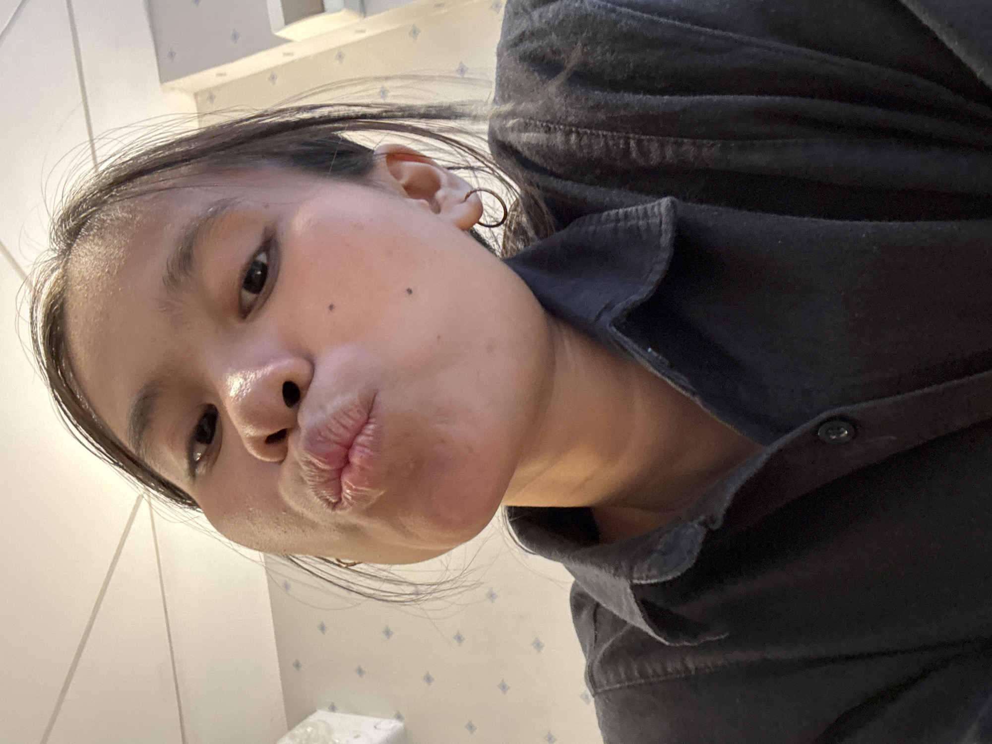 A young woman, facing forward and making a kissing face, is captured in a close-up, overhead shot. She is wearing a dark collared shirt. A small, circular hoop earring is visible on her right ear. The background appears to be an indoor setting with a patterned wallpaper and a textured ceiling. The image is a selfie taken from a low angle, with the woman's face filling most of the frame. The lighting is bright and appears to be natural.