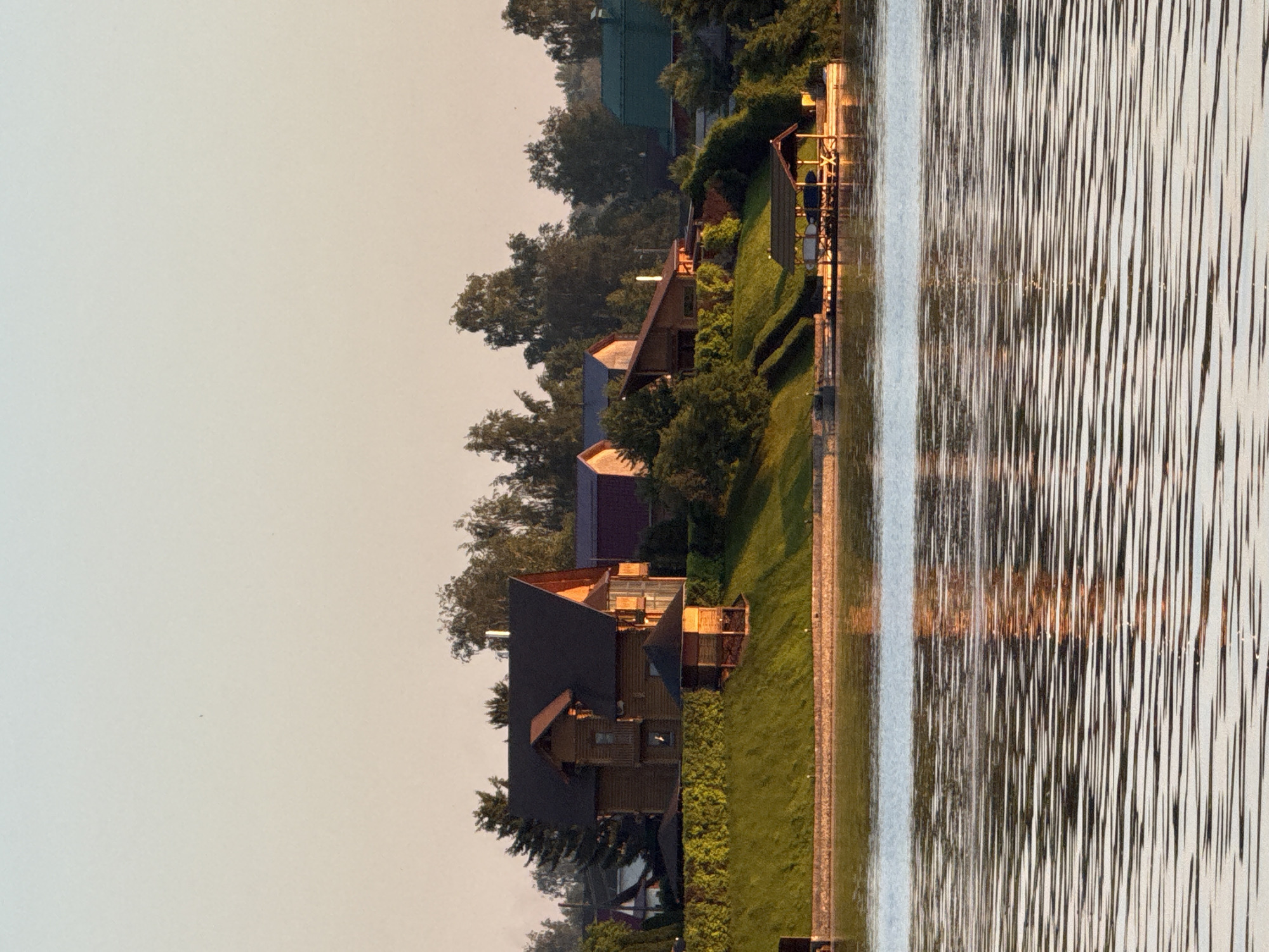 This image depicts a waterside landscape in what is described as Votkinsk, Russia, captured during either dawn or dusk. The upper portion of the frame shows a clear sky with a subtle gradient, transitioning from a light blue at the zenith to a pale orange-yellow closer to the horizon.
The foreground is dominated by a body of water, likely a river or lake, exhibiting numerous horizontal ripples that create elongated and distorted reflections of the distant shoreline and sky.
The middle ground features a gently sloping landmass or bank, covered with neatly maintained green grass and dense green foliage, possibly hedges or bushes, particularly in front of the primary structures. A stone or concrete retaining wall forms the immediate edge between the land and the water.
Prominently situated on the grassy slope is a two-story wooden house with a dark, multi-gabled roof and light brown wooden siding. A small, open-sided wooden gazebo with a dark roof stands adjacent to this house on the slope. Further to the right, several other houses are visible amidst trees, including one with a distinct green roof and others with darker roofs. Various deciduous and coniferous trees dot the landscape behind and around the houses.
Extending into the water from the right side of the bank is a wooden dock featuring a covered boat shed with a dark roof. Two small, light-colored boats, appearing to be kayaks or canoes, are stored underneath the shed's cover.
No human figures are visible in the scene. The overall lighting is soft and warm, indicative of early morning or late afternoon conditions.