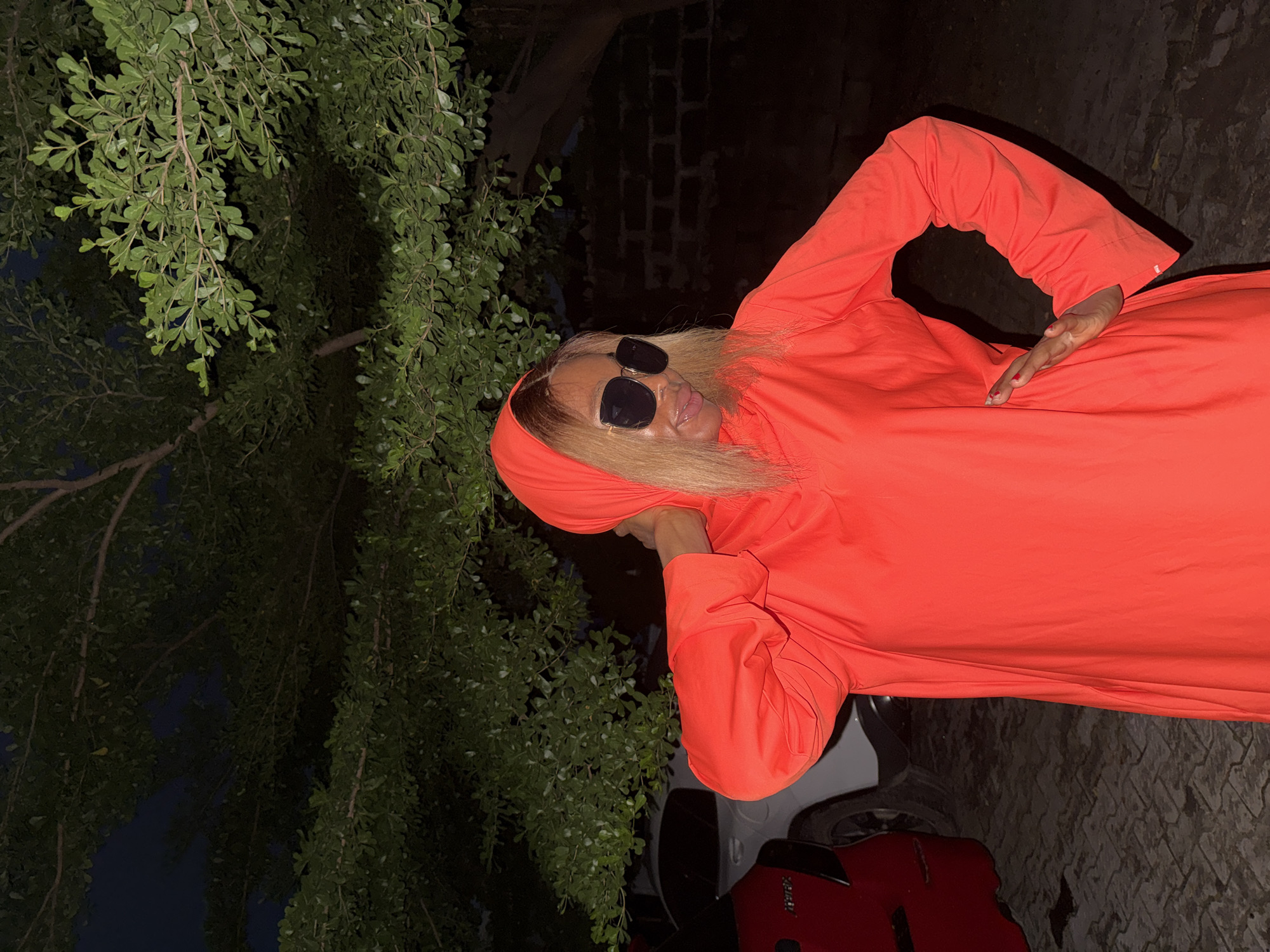 An individual stands outdoors at night in Ado, Nigeria. The person is wearing a bright orange hooded garment and large sunglasses, with their hair blonde and shoulder-length. They are posing with one hand on their neck and the other on their hip. Behind them, a red car is visible on the left, and a white car is partially visible to the right of that. Trees with green foliage are overhead, and a dark brick wall forms the background on the right. The ground appears to be paved with stone.