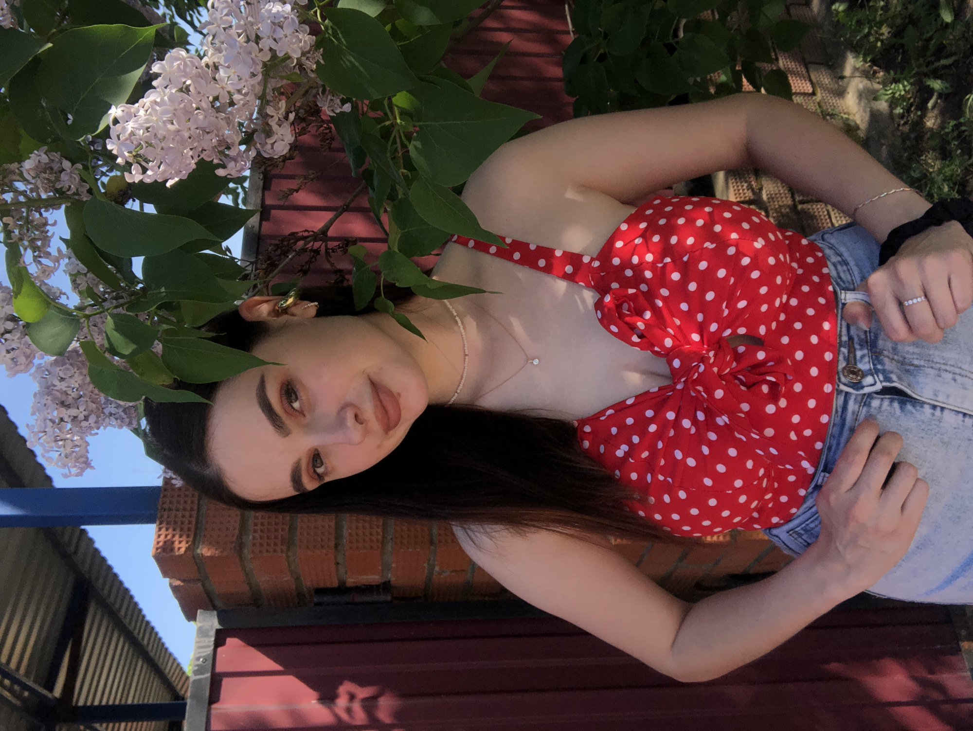 A young woman stands in front of a brick wall and fence, positioned in a slightly angled shot. She has long, dark brown hair and fair skin, wearing a red polka-dot top and light blue jeans. Her hand rests on the jeans near the waistband, and she wears a ring on her finger. A necklace is visible around her neck. To her left, lilac blossoms frame the shot, obscuring part of the wall. The setting suggests an outdoor location, likely in a residential or park area, in the city of Noviy, Russia. A corner of a building roof is visible in the background, implying the vicinity of structures.