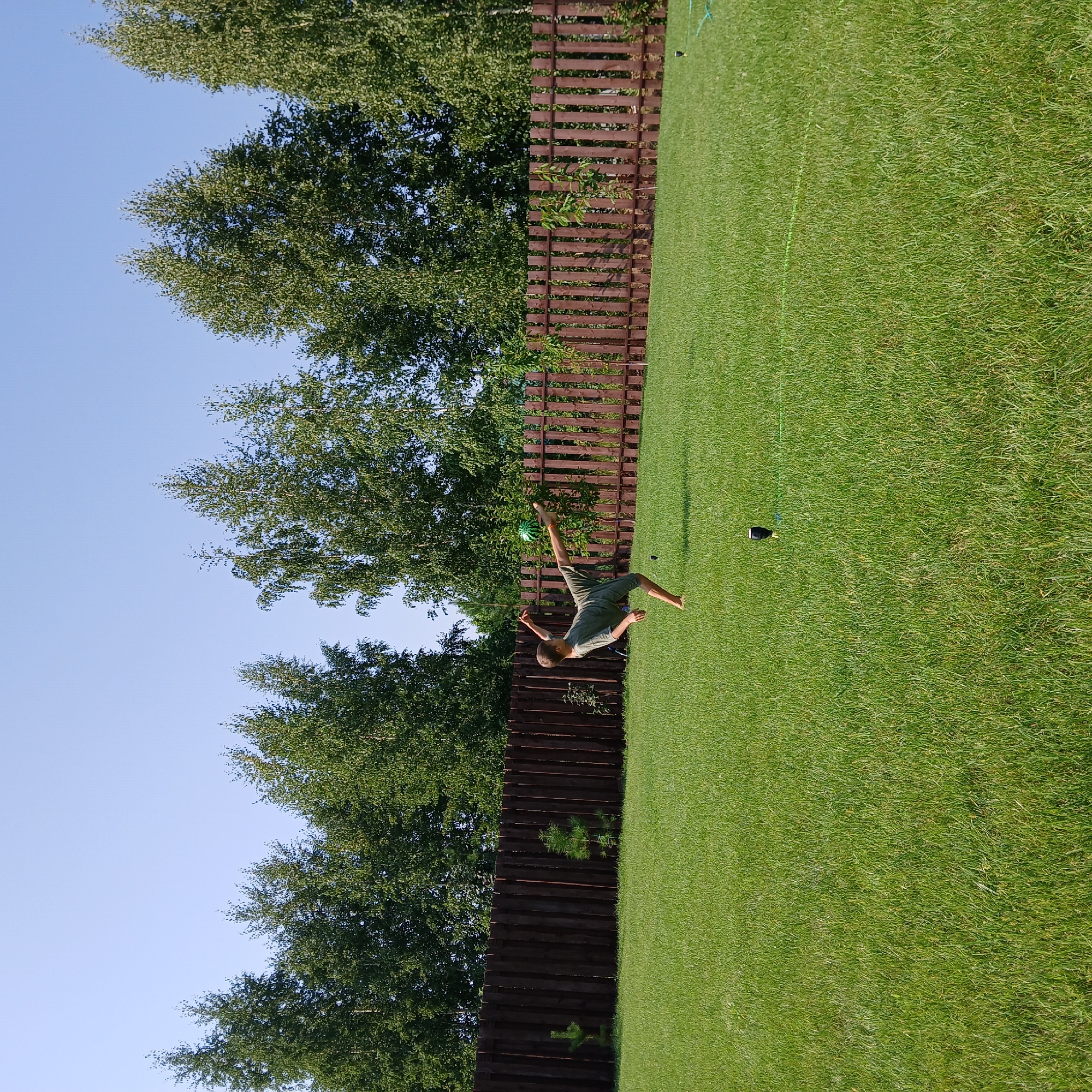 In a sunlit backyard in Samarsky, Russia, a young boy is captured mid-air, performing a bicycle kick to a green and white soccer ball. He wears a green t-shirt and shorts, with bare feet. The boy is positioned on a lush, green lawn that extends towards a tall, dark brown wooden fence. Behind the fence, a line of mature trees with vibrant green foliage forms a natural backdrop. The sky is a clear, bright blue, indicating a summer day. A black object, possibly a toy or piece of equipment, is visible on the grass in the foreground, and a green hose or string is laid out on the lawn.