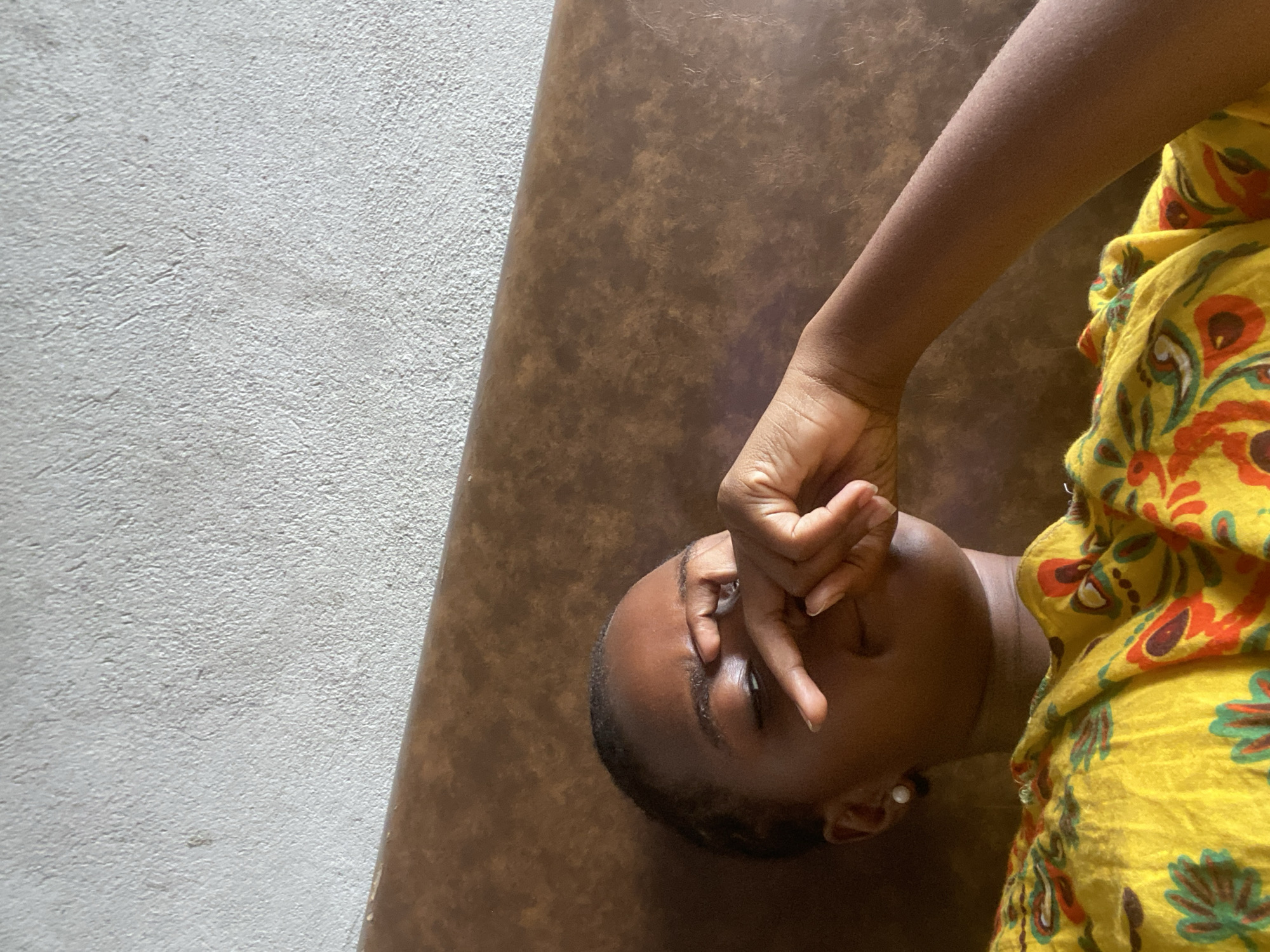 A woman lies on a brown surface, possibly a mat or table, against a light gray wall. She has dark skin and short, dark hair. Her right hand is raised to her face, partially covering her eyes. She wears a yellow top with a floral pattern in red, green, and brown. The environment is a building. There is no information available within the image to confirm it is Dar es-Salaam, Tanzania.